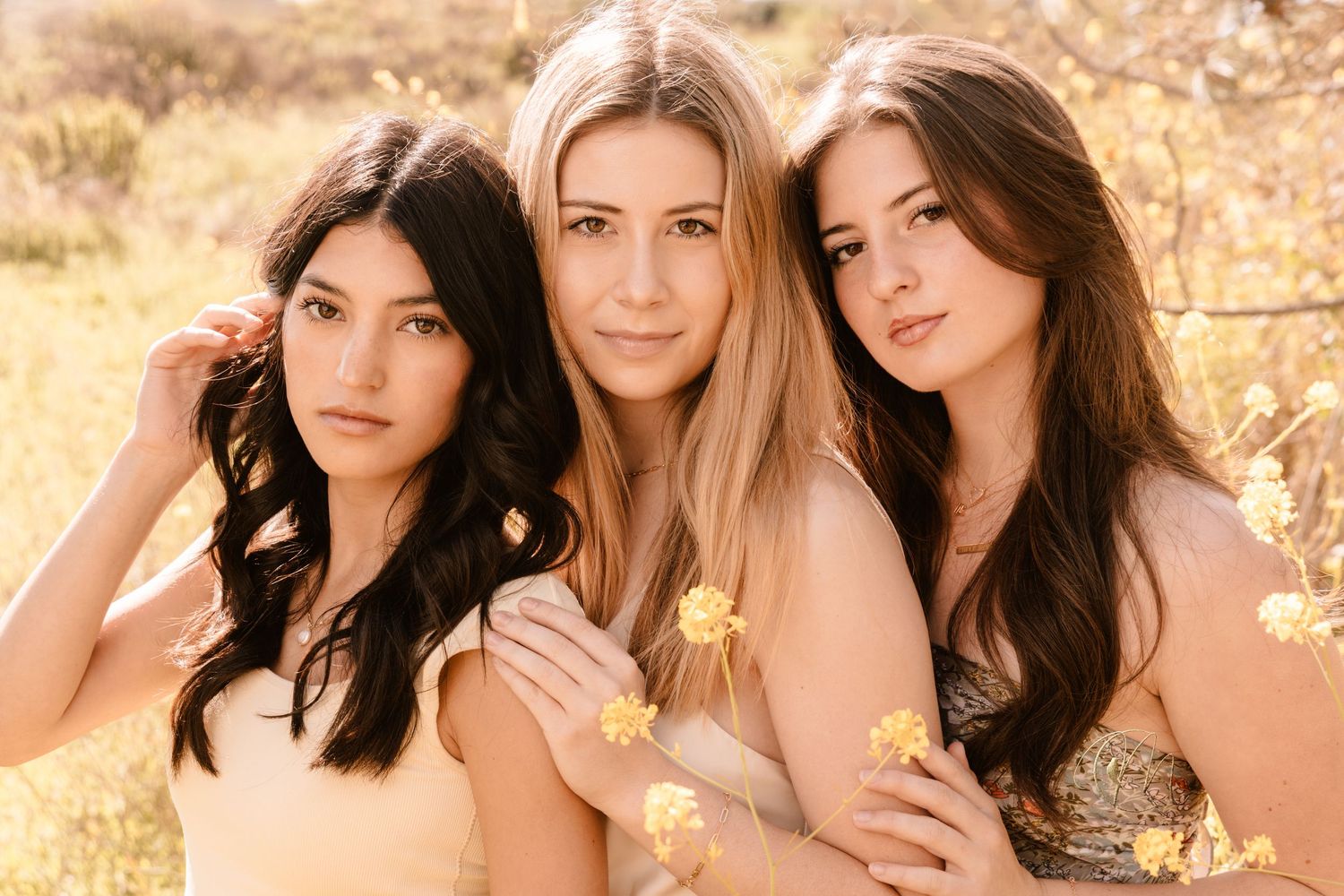 Three young women with long hair pose together outdoors among golden wildflowers in warm sunlight.
