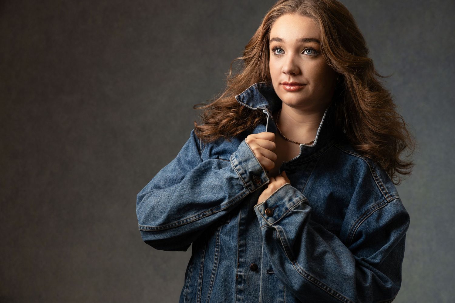 Young woman with wavy auburn hair wearing an oversized denim jacket, posing against a gray studio background.