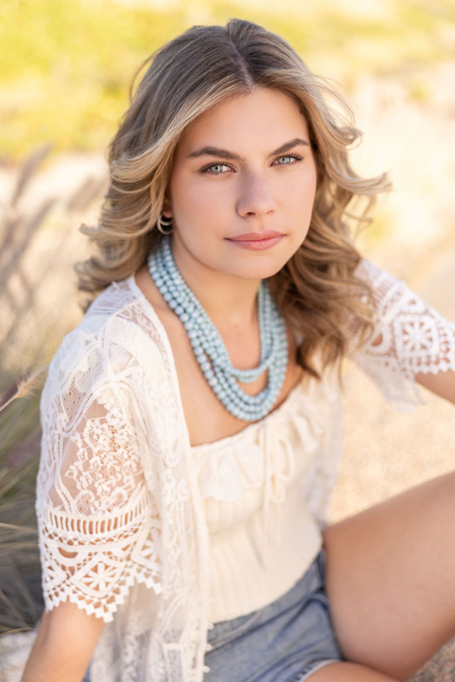 Young woman with wavy blonde hair wearing a white lace top and turquoise beaded necklace, posing outdoors in warm light.