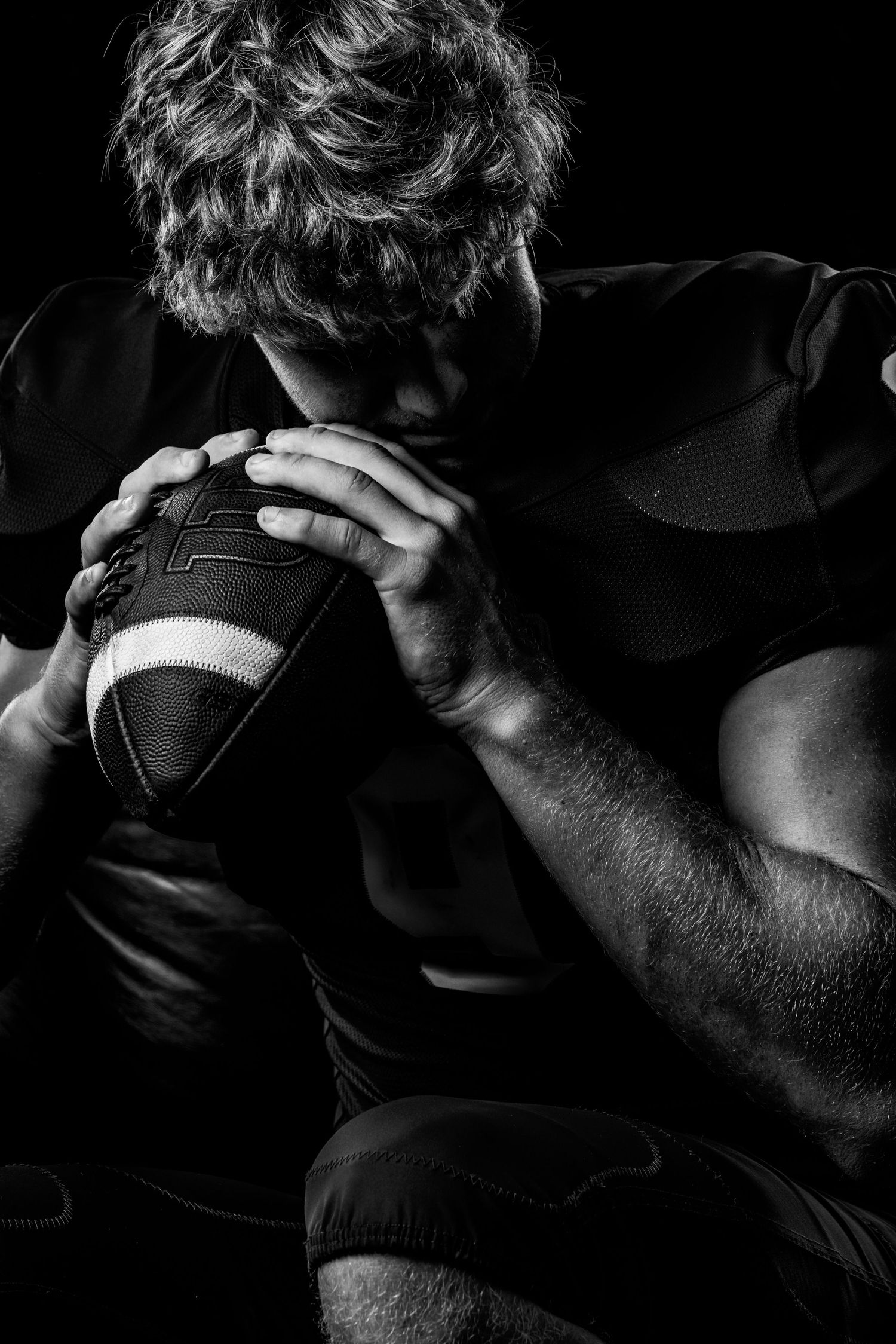 Black and white photo of a focused American football player holding a football close to his face in dramatic lighting.