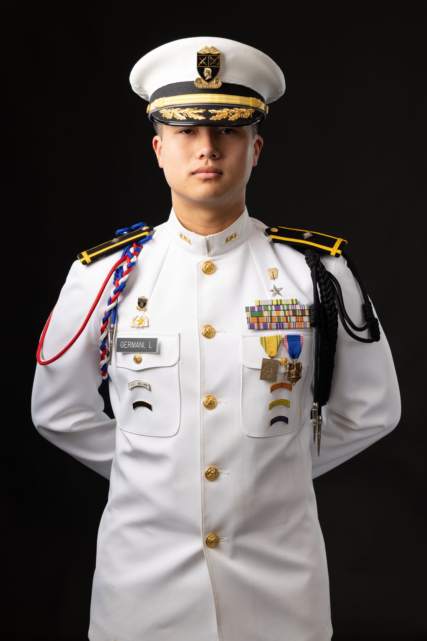Young military officer in white dress uniform with medals, epaulettes, and white cap poses against dark background.