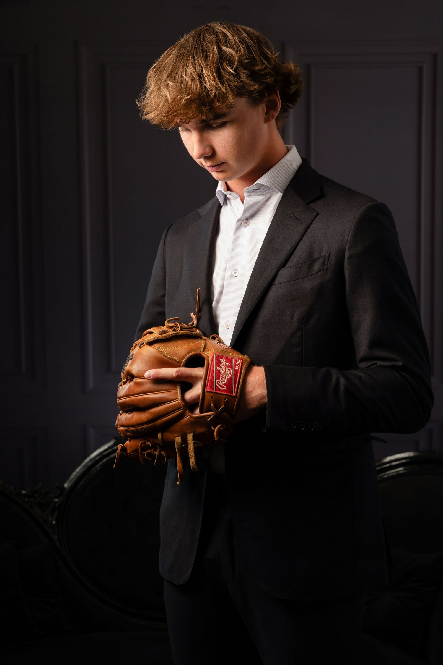 Young man in black suit holding a Rawlings baseball glove, posing against a dark gray wall.
