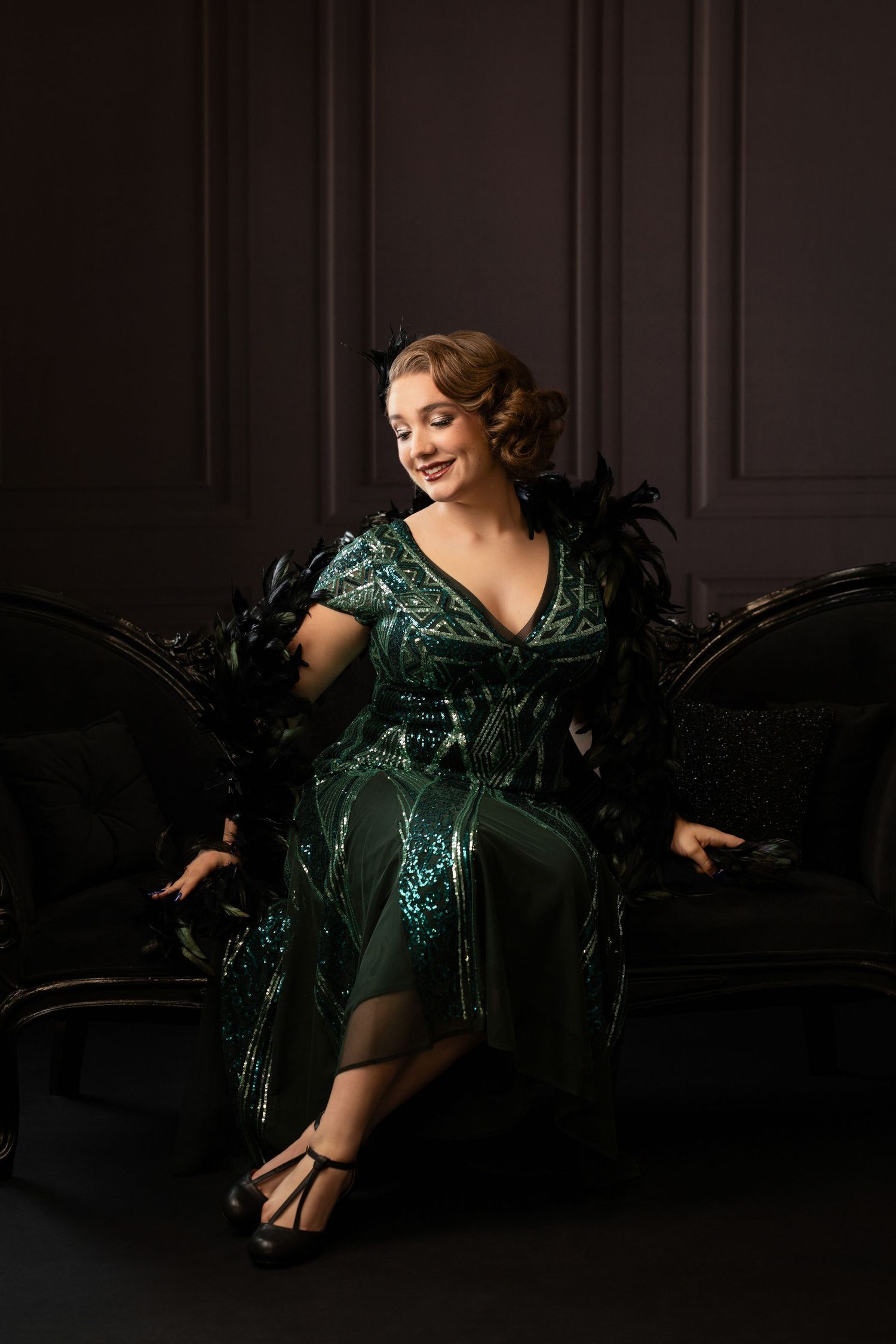 Woman in green sequin flapper dress with black feathers sits smiling on dark velvet sofa.