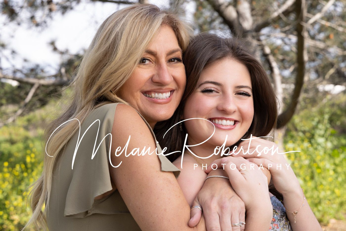 Two smiling women embrace outdoors among yellow wildflowers in a warm portrait by Melanie Robertson Photography.
