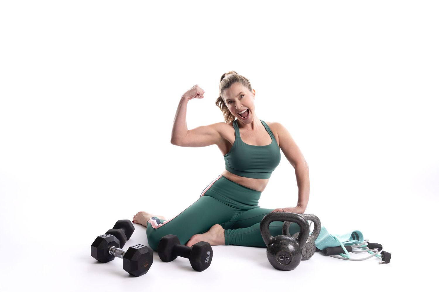 Fit woman in green activewear flexing bicep while sitting with dumbbells and kettlebell on white background.