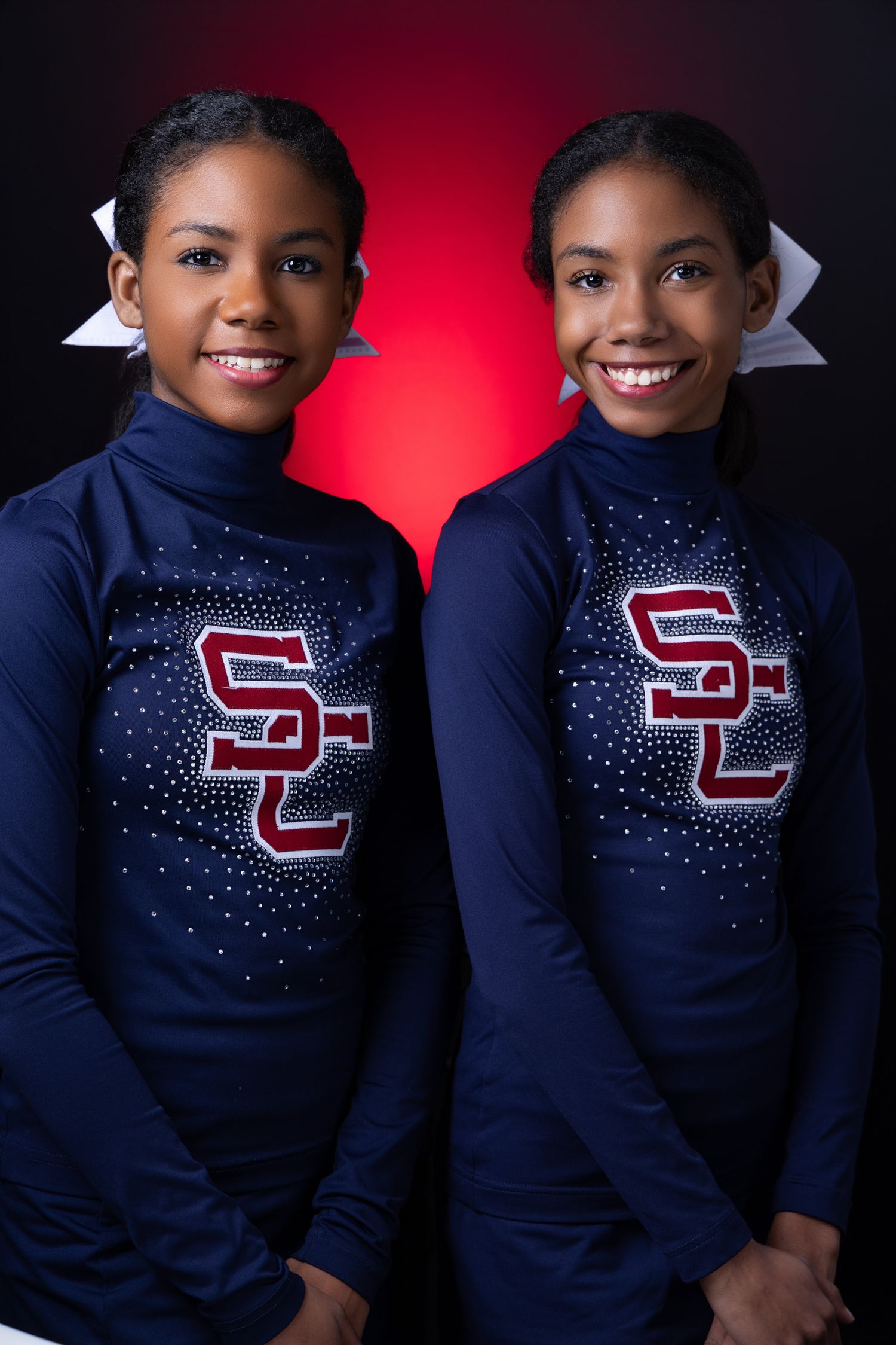 Two young girls in matching navy SC cheerleading uniforms smiling against a red and black background.