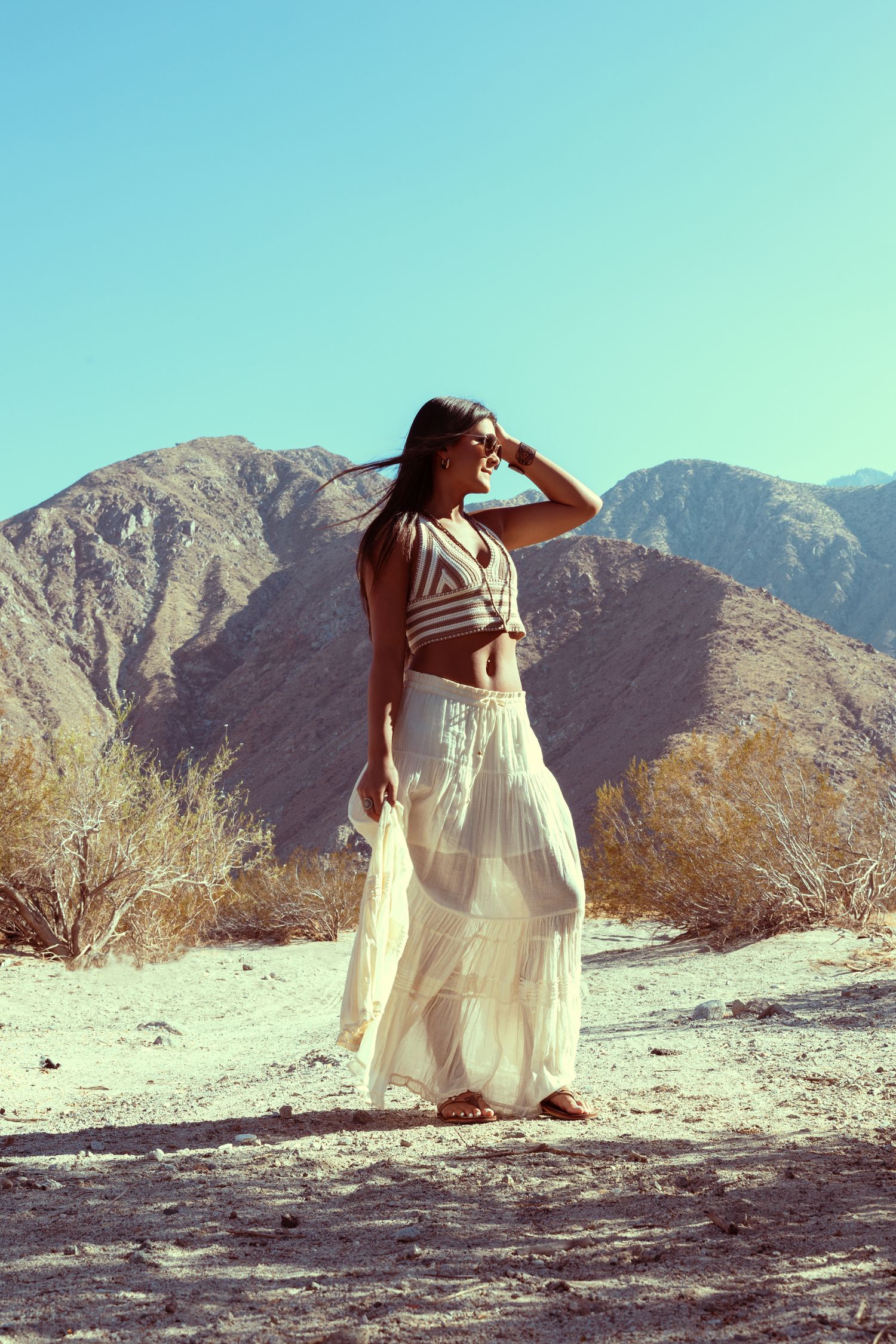 Woman in white maxi skirt and crop top posing in desert landscape with mountains and clear blue sky.