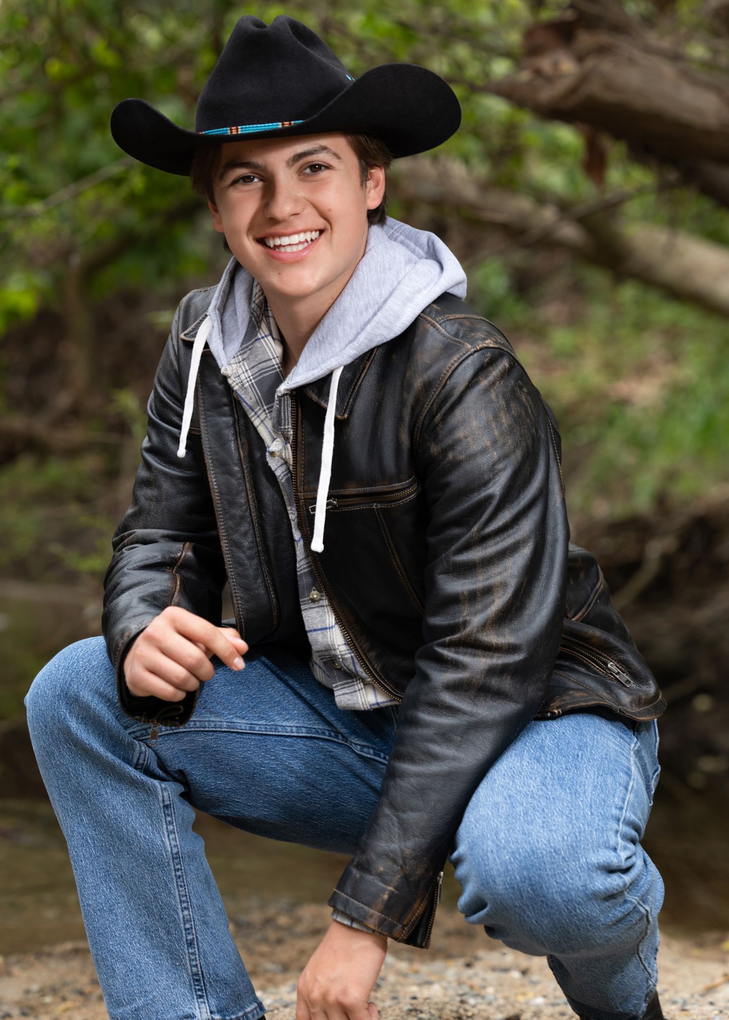 Smiling young man in black cowboy hat and leather jacket crouching outdoors near a creek.
