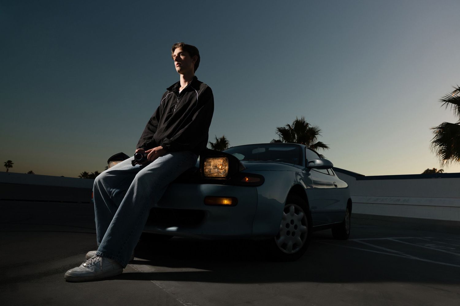 Man leans on car hood at sunset with palm trees in background.