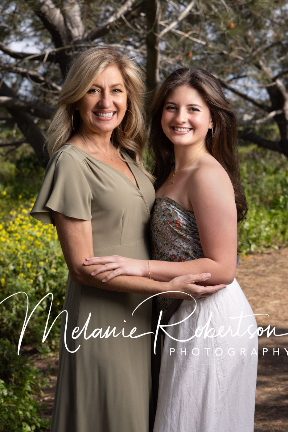 Two smiling women pose outdoors among trees and wildflowers in a professional portrait by Melanie Robertson Photography.