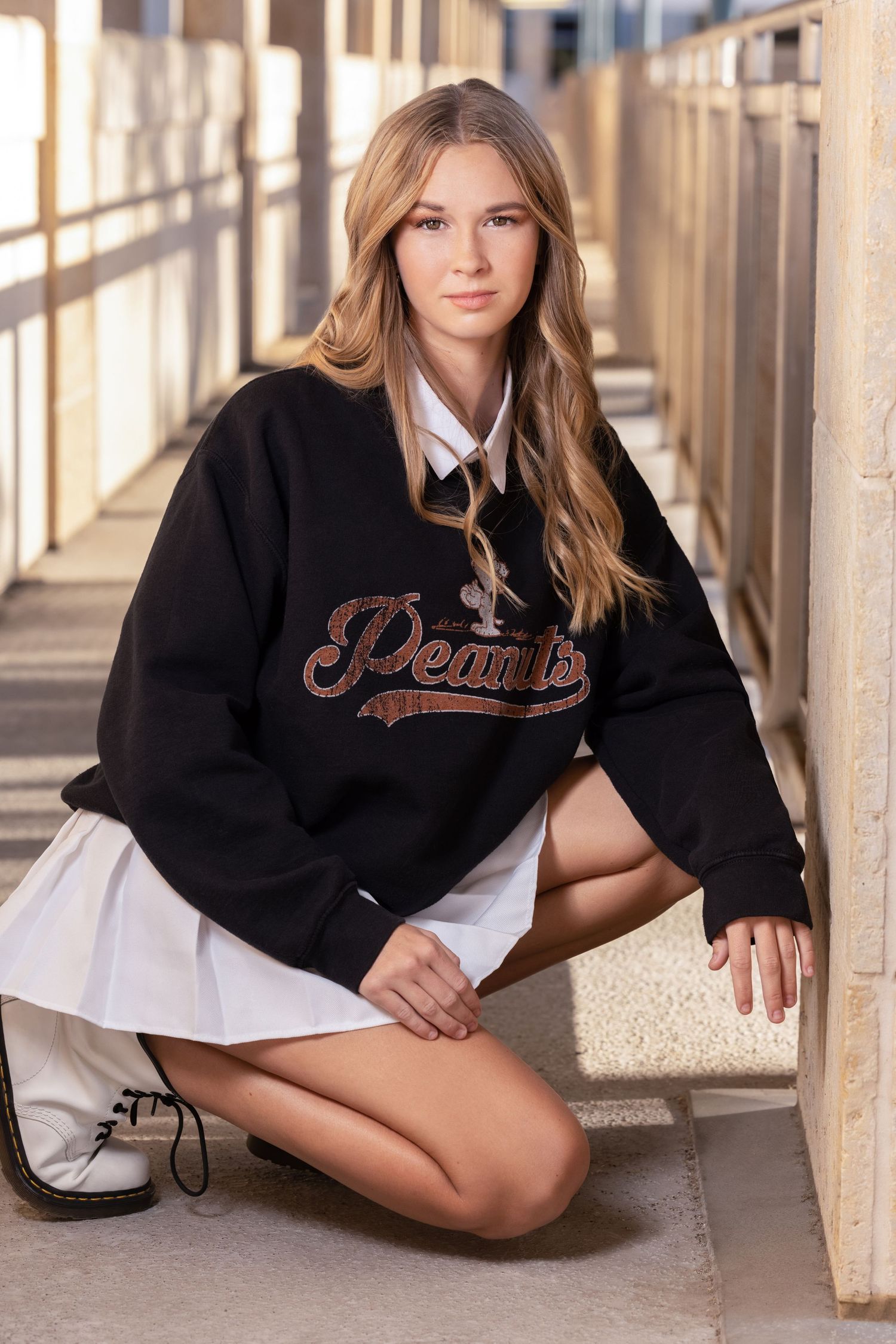 Young woman in black Peanuts sweatshirt and white pleated skirt kneeling outdoors in a sunlit corridor.