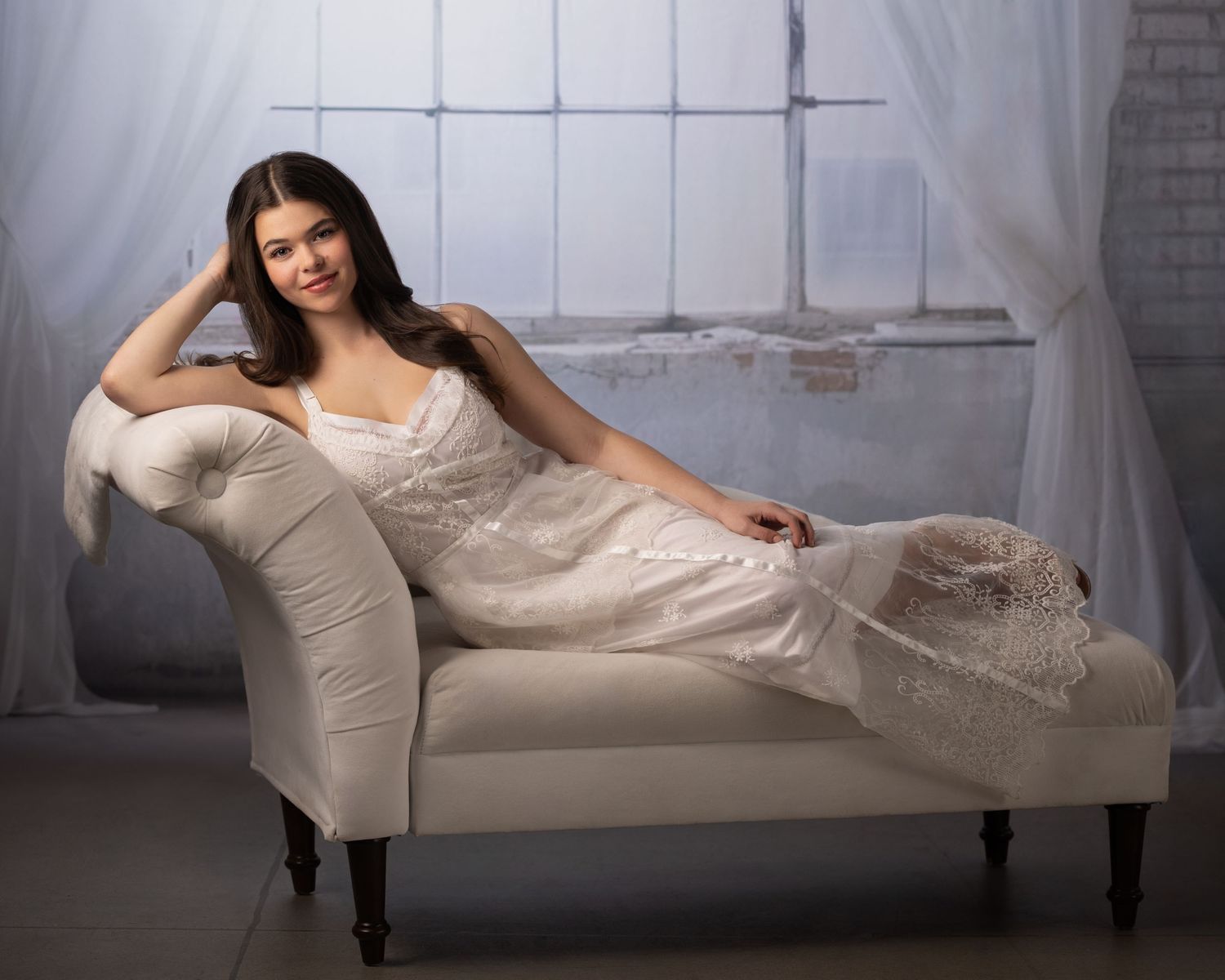 Young woman in white lace gown reclines gracefully on ivory chaise lounge before sheer curtained window.