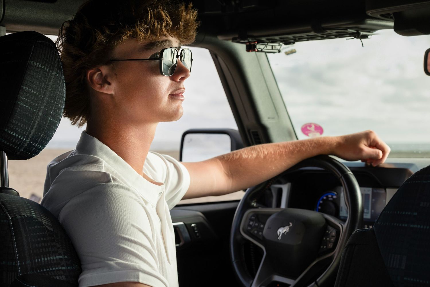 Young man with sunglasses driving a car, hand on steering wheel, wearing a white polo shirt.