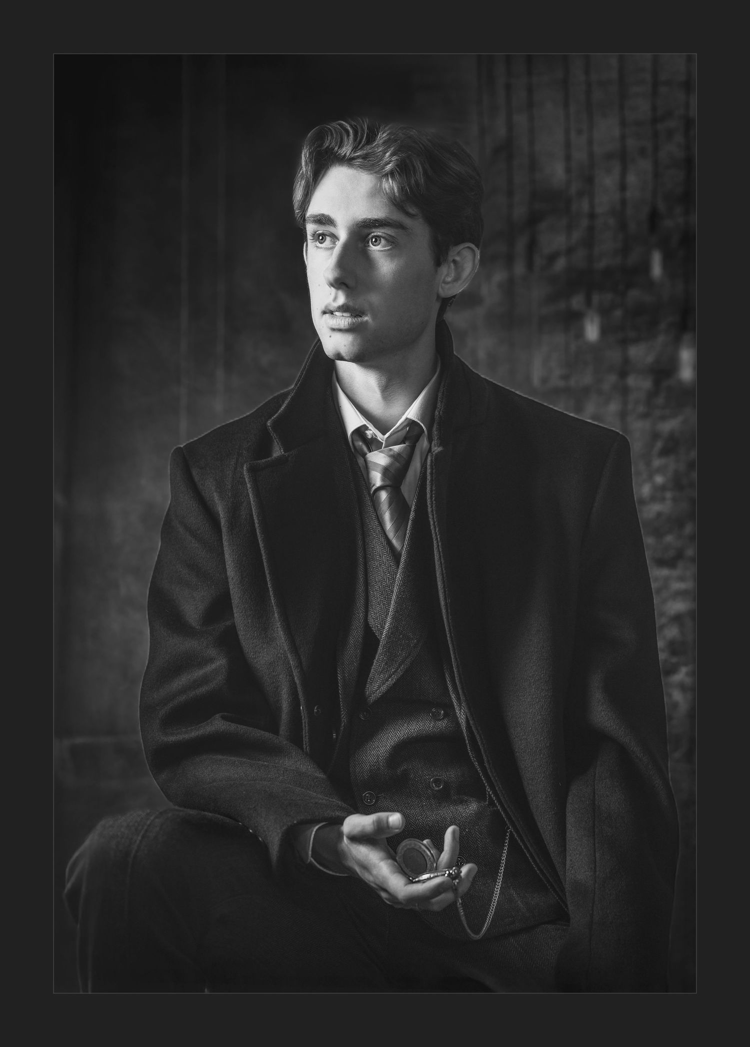 Black and white portrait of a young man in a vintage suit and overcoat, seated and gazing thoughtfully to the side.