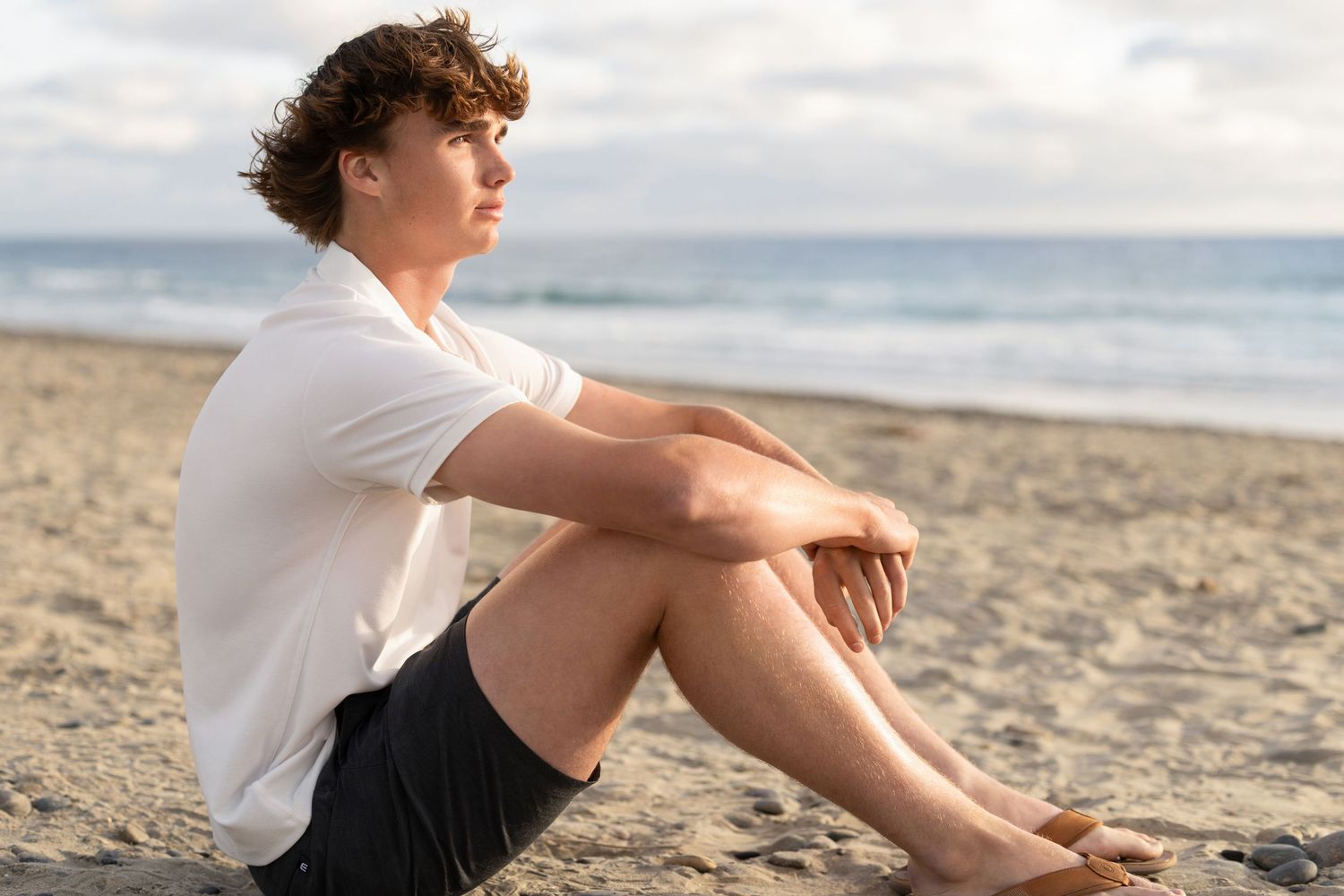 Young man with curly hair in a white shirt and dark shorts sits on a sandy beach, gazing thoughtfully toward the ocean at sunset.