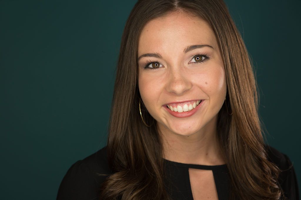 Professional headshot of smiling young brunette woman in black top against teal background.