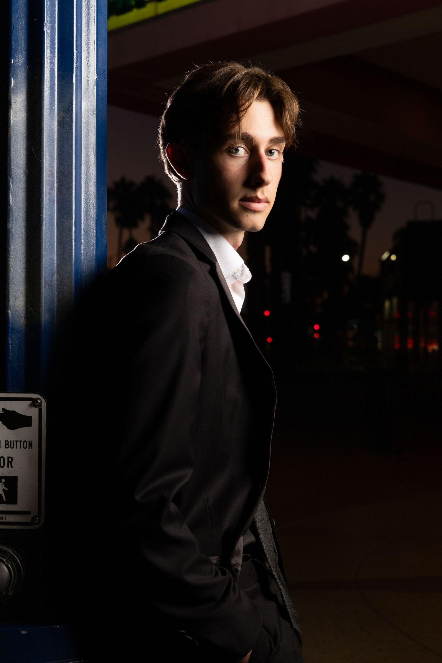 Young man in black suit leaning against blue pole at night with city lights in background.