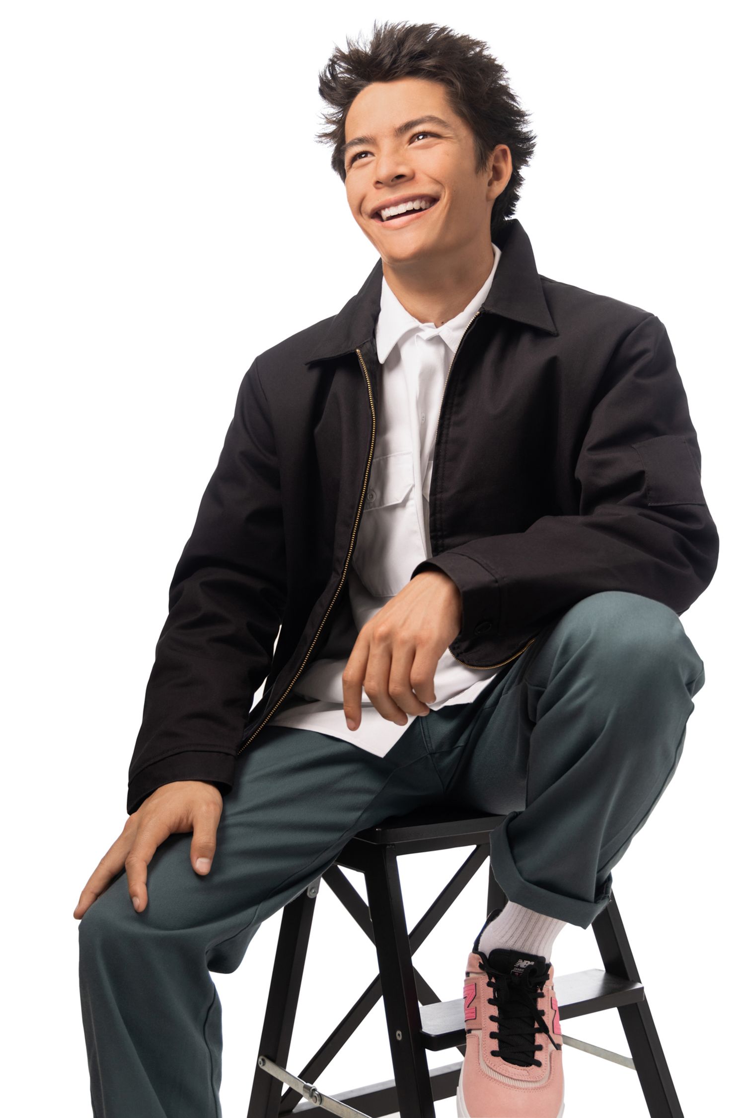 Smiling young man in black jacket, green pants, and pink sneakers sitting on a stool against white background.