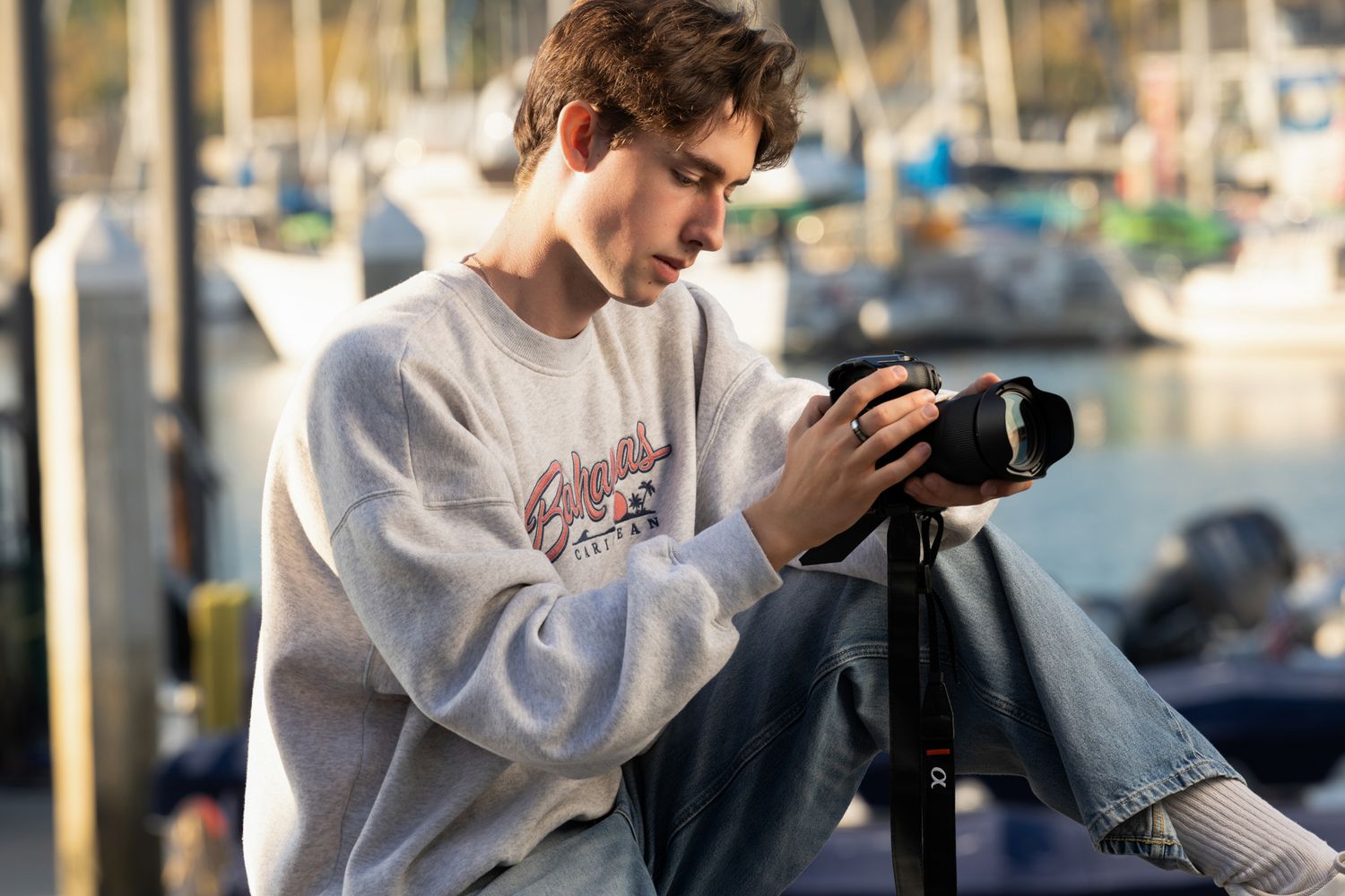 Young man in gray sweatshirt reviewing photos on a DSLR camera at a marina.