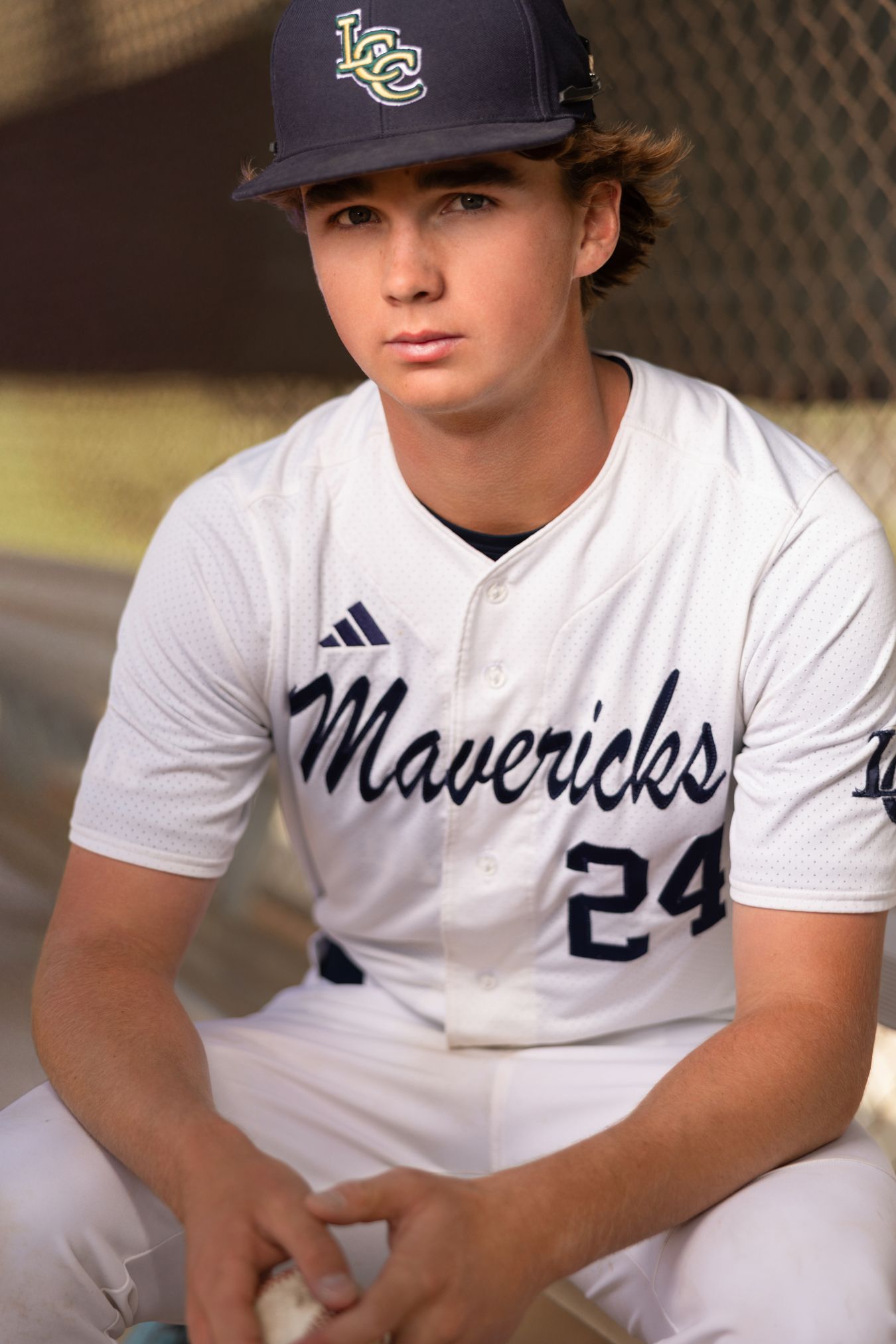 Young baseball player in Mavericks #24 white uniform and navy cap sits in dugout holding a baseball.