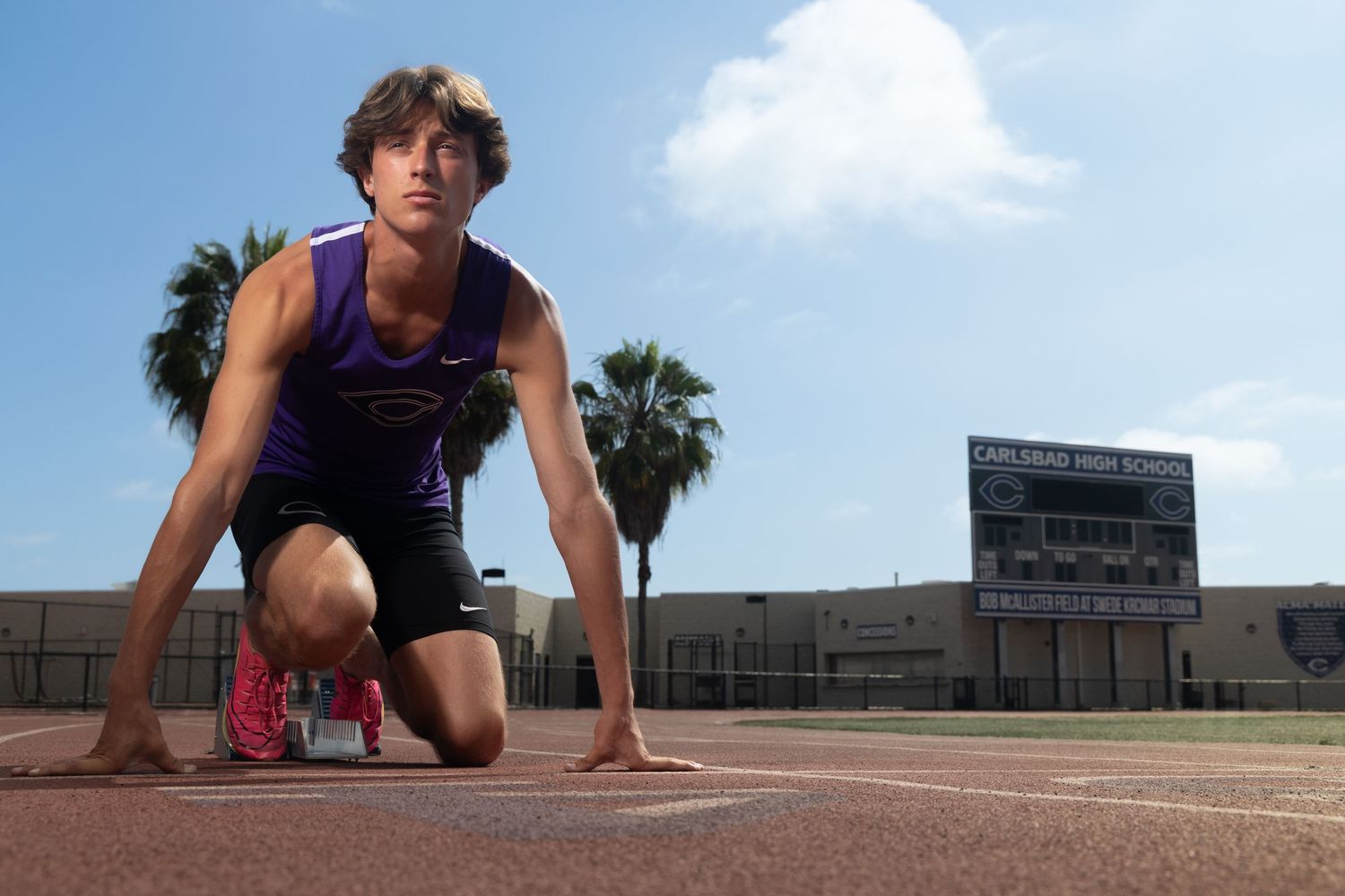 Male sprinter in a purple jersey crouches in the starting position on a red track with palm trees and a high school scoreboard behind him.