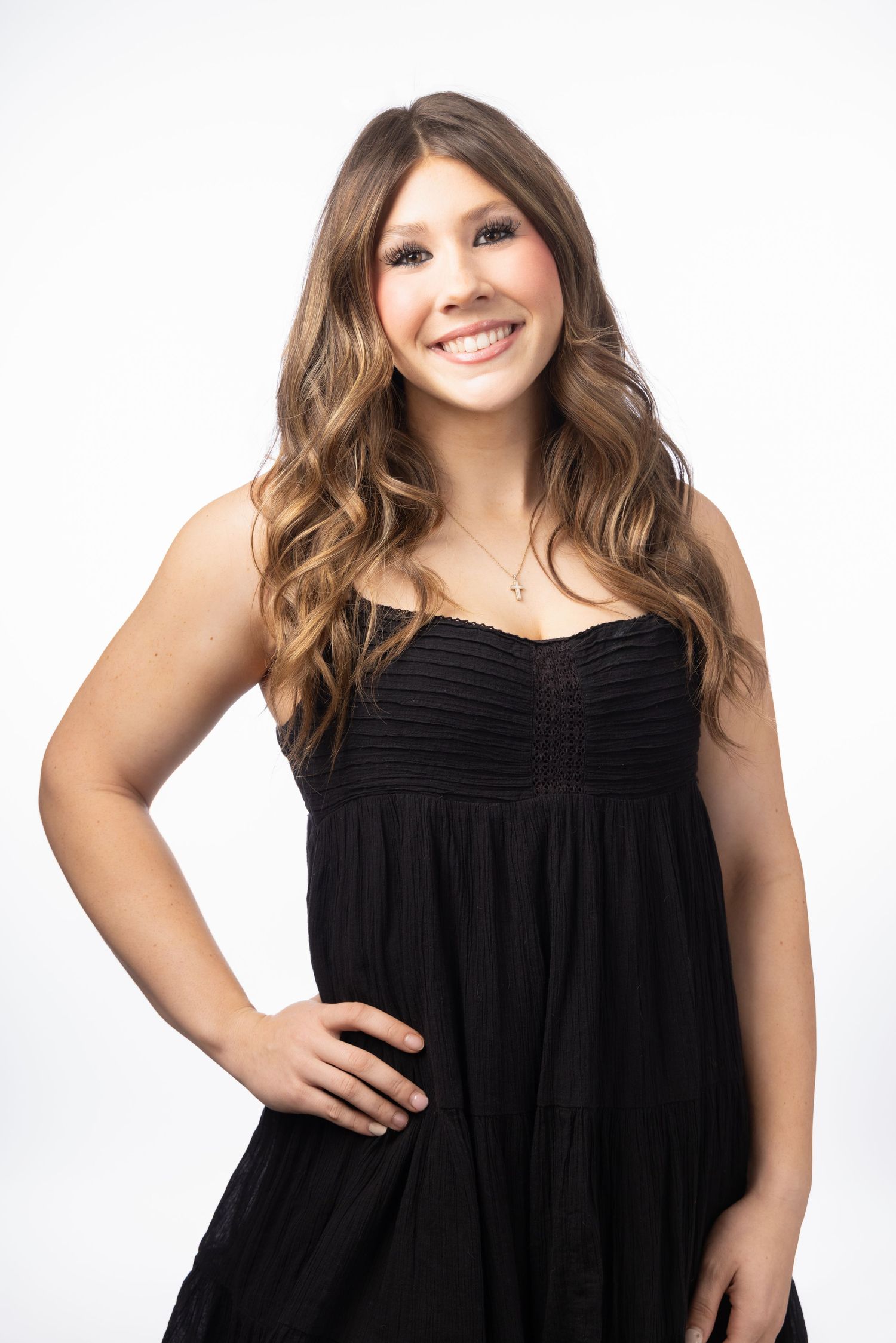 Young woman with long wavy brown hair smiling in a black strapless dress and necklace against white background.