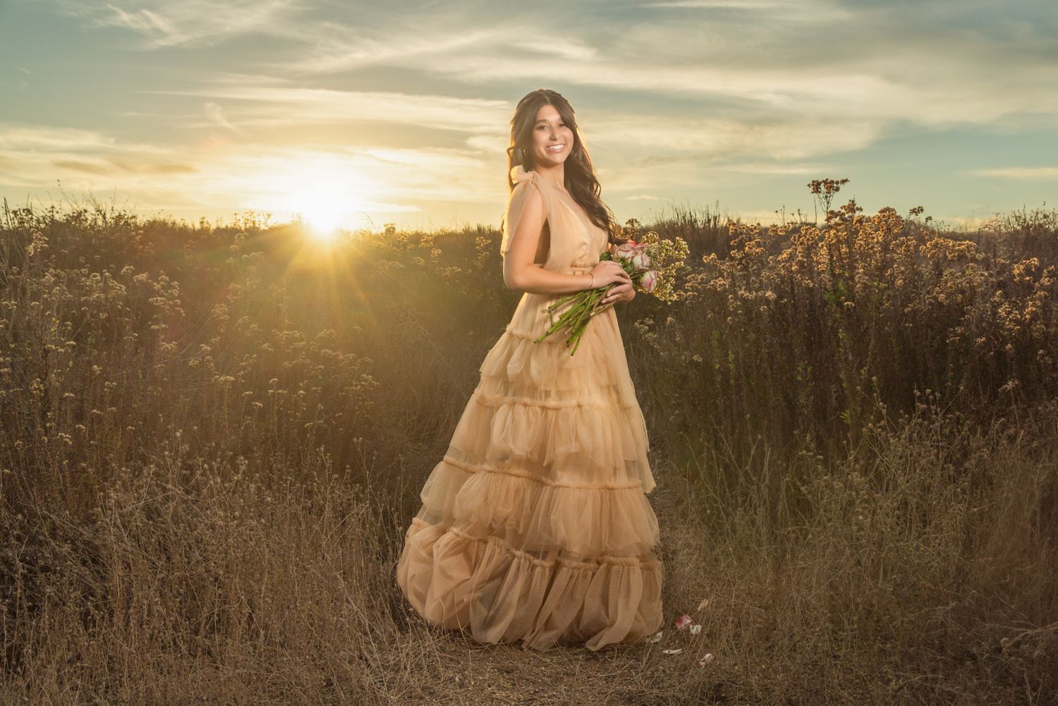 Woman in golden tiered gown holding wildflowers in a sunlit field at sunset.