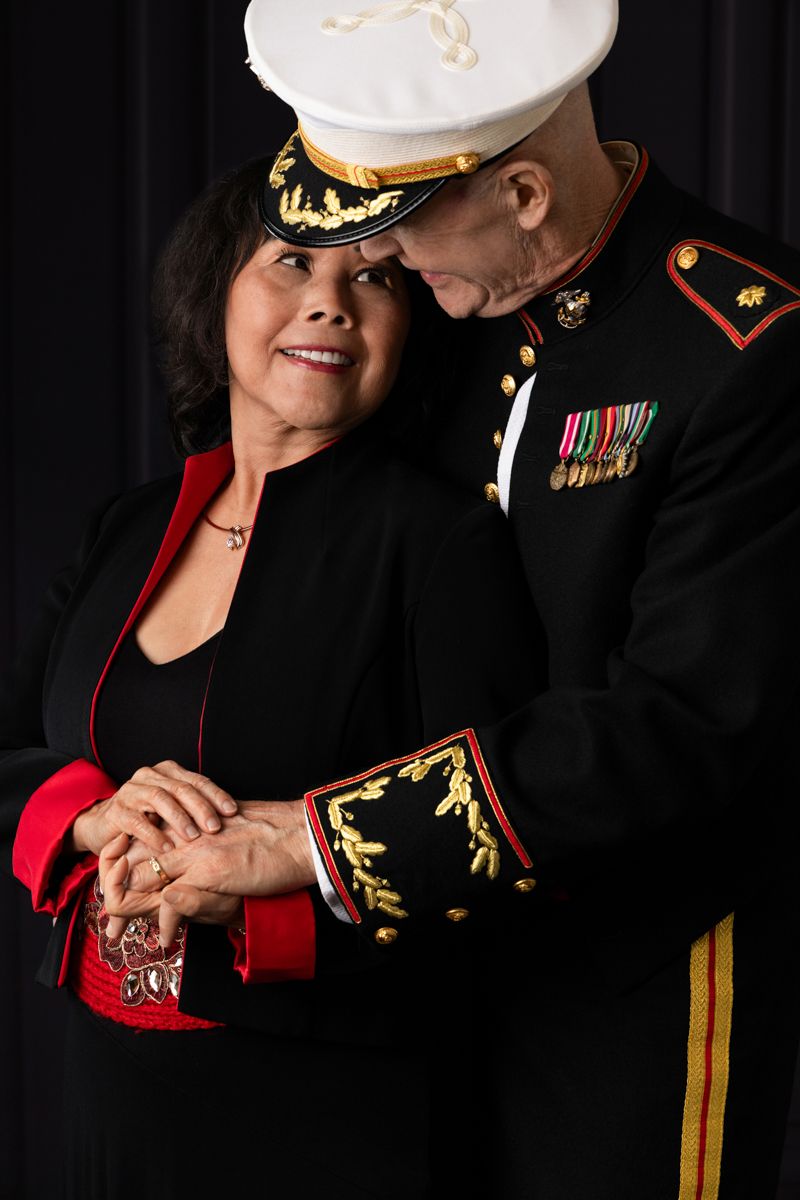 A Marine in dress uniform tenderly embraces a smiling woman in black and red attire against a dark background.