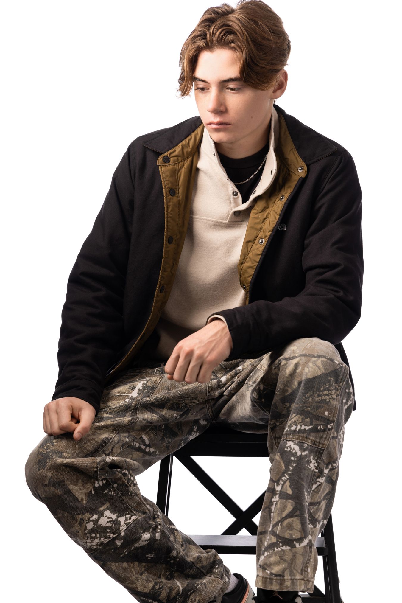 Young man sitting on stool wearing black jacket, camo pants, and layered tops in studio portrait.