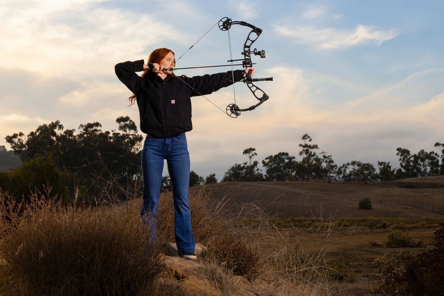Woman aiming compound bow outdoors at sunset in rural landscape, wearing black hoodie and jeans.