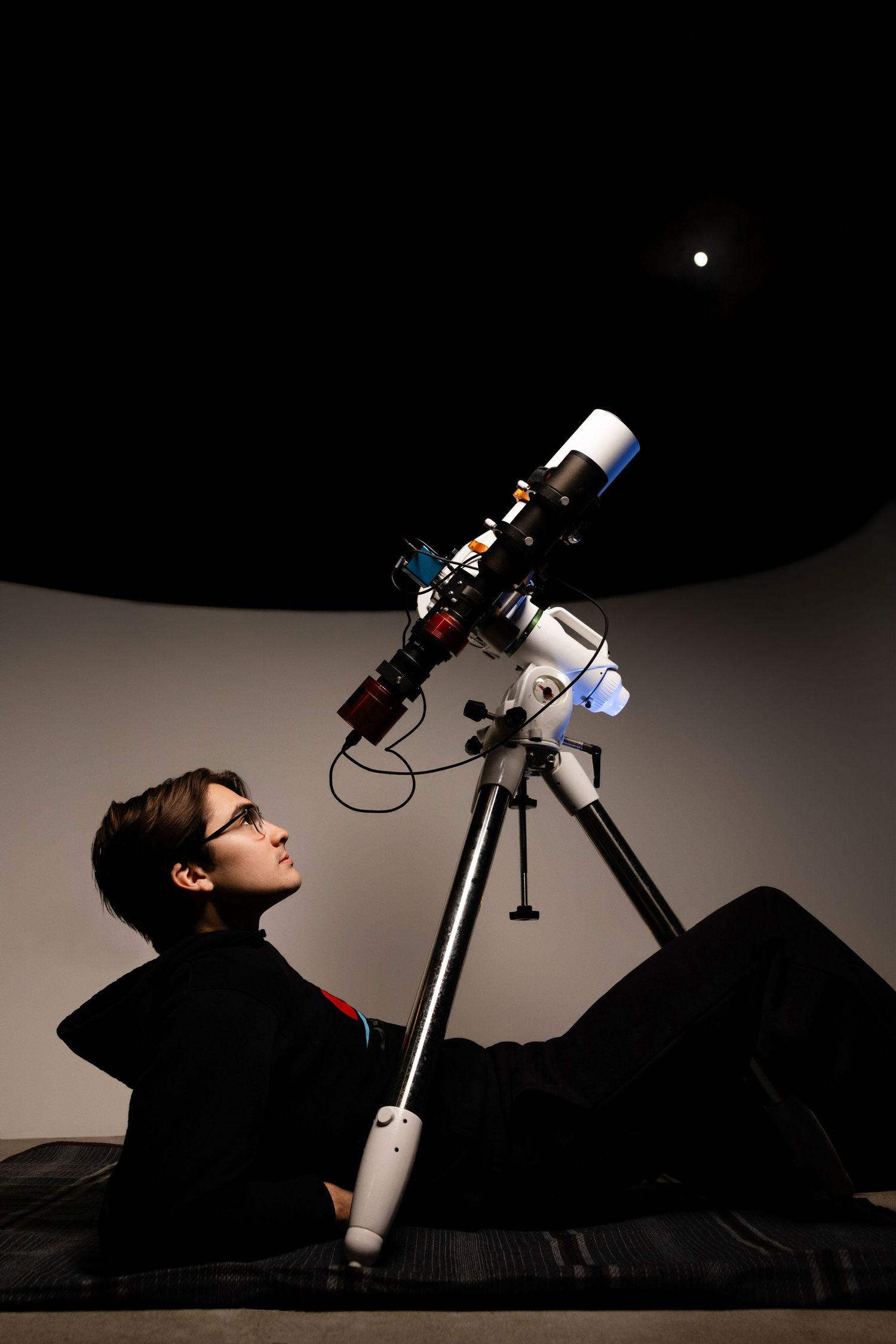 Person lying on floor gazing up at telescope on tripod in dark planetarium with glowing light on ceiling.