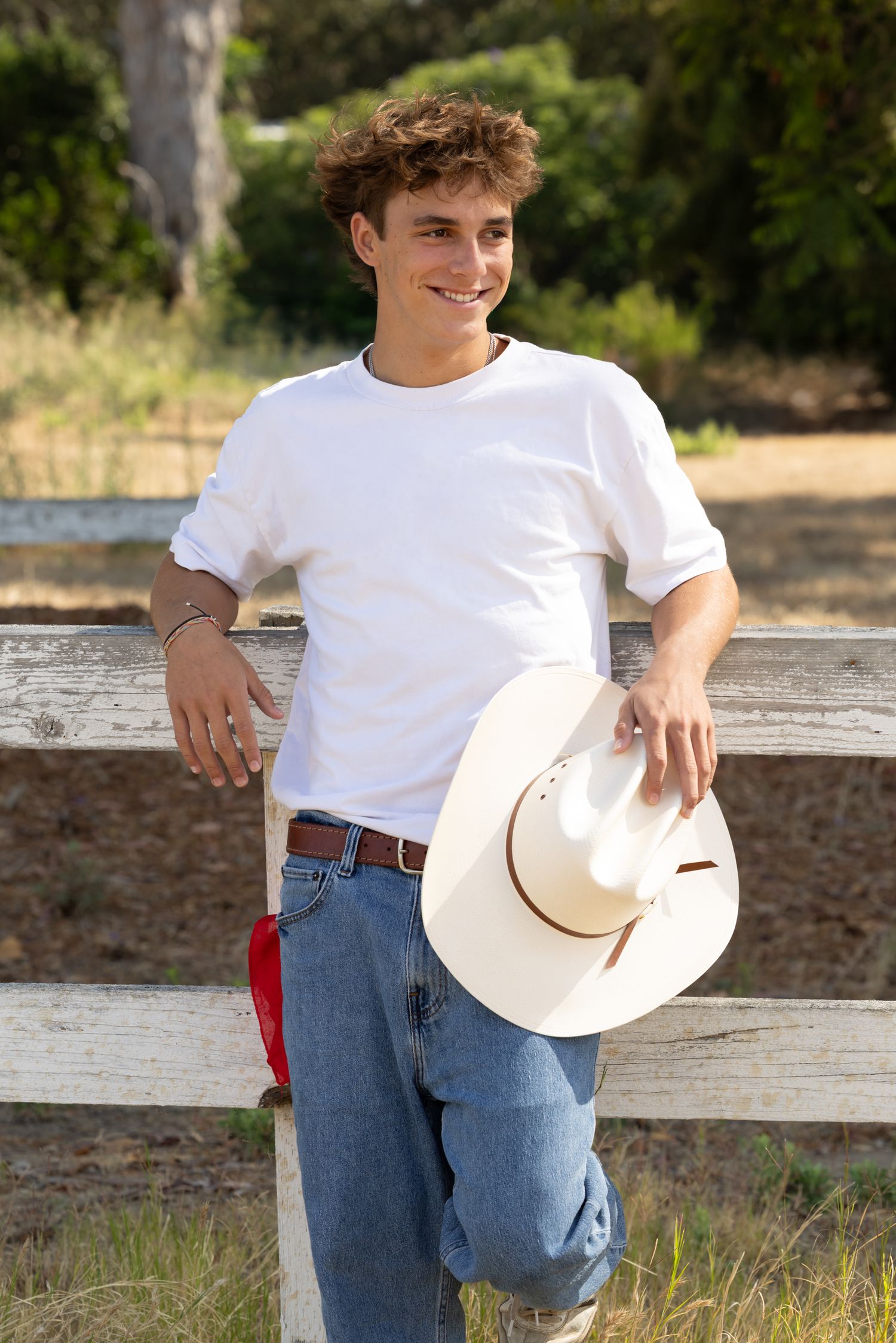 Young man in white t-shirt and jeans holding a cowboy hat, leaning on a wooden fence outdoors.