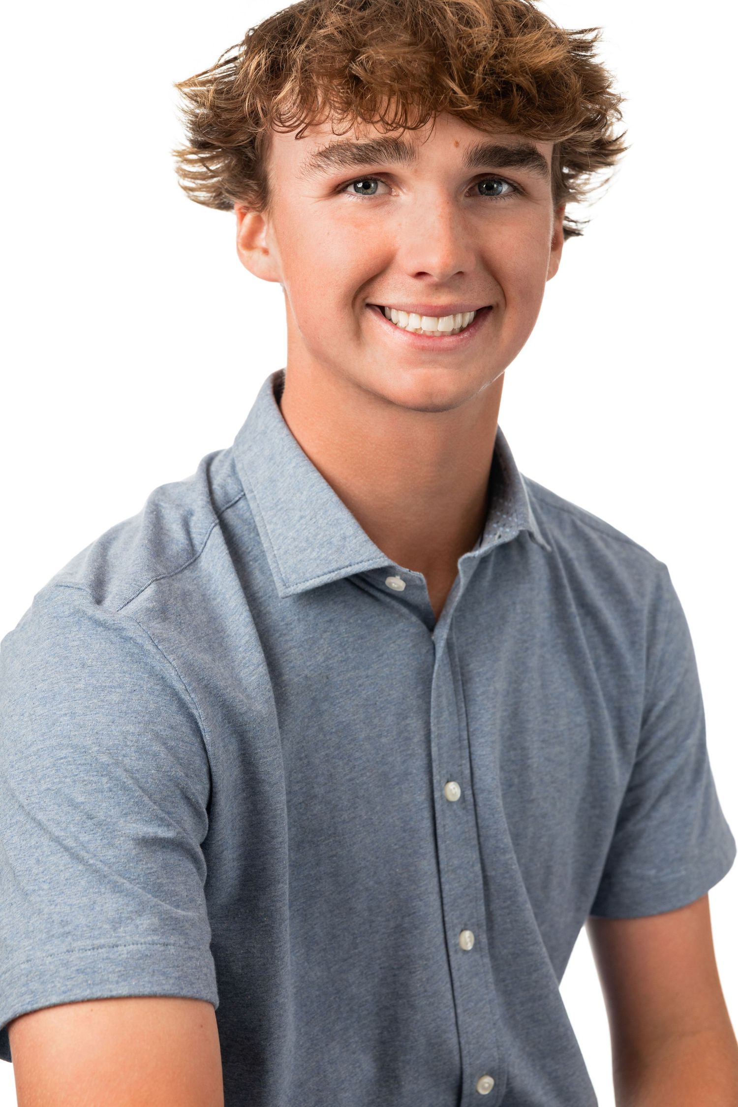 Smiling teenage boy with curly brown hair wearing a gray polo shirt against a white background.
