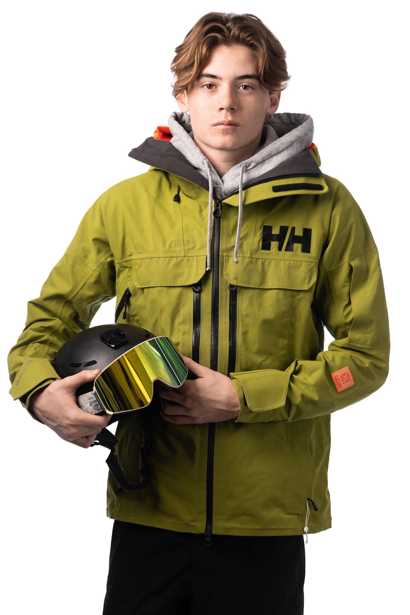 Young man wearing an olive green Helly Hansen ski jacket, holding a black helmet with yellow-tinted goggles.