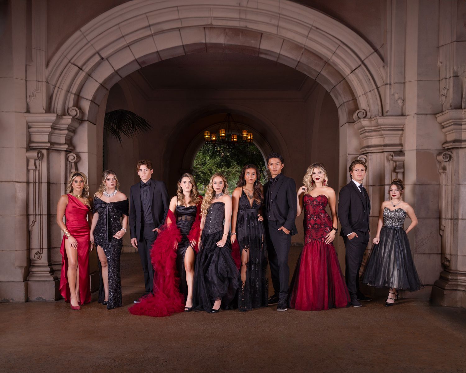 Group of elegantly dressed teens posing in formal gowns and suits under a stone archway for prom photos.