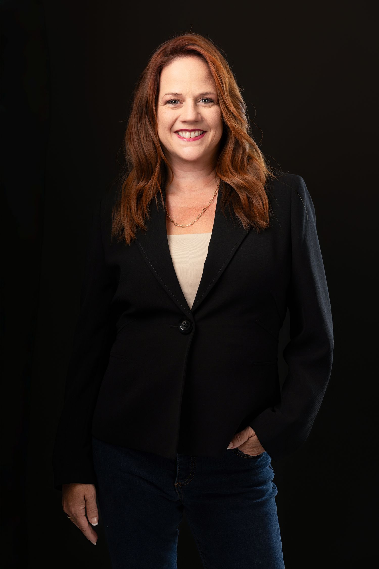 A smiling woman with red hair wearing a black blazer and jeans poses confidently against a dark studio background.