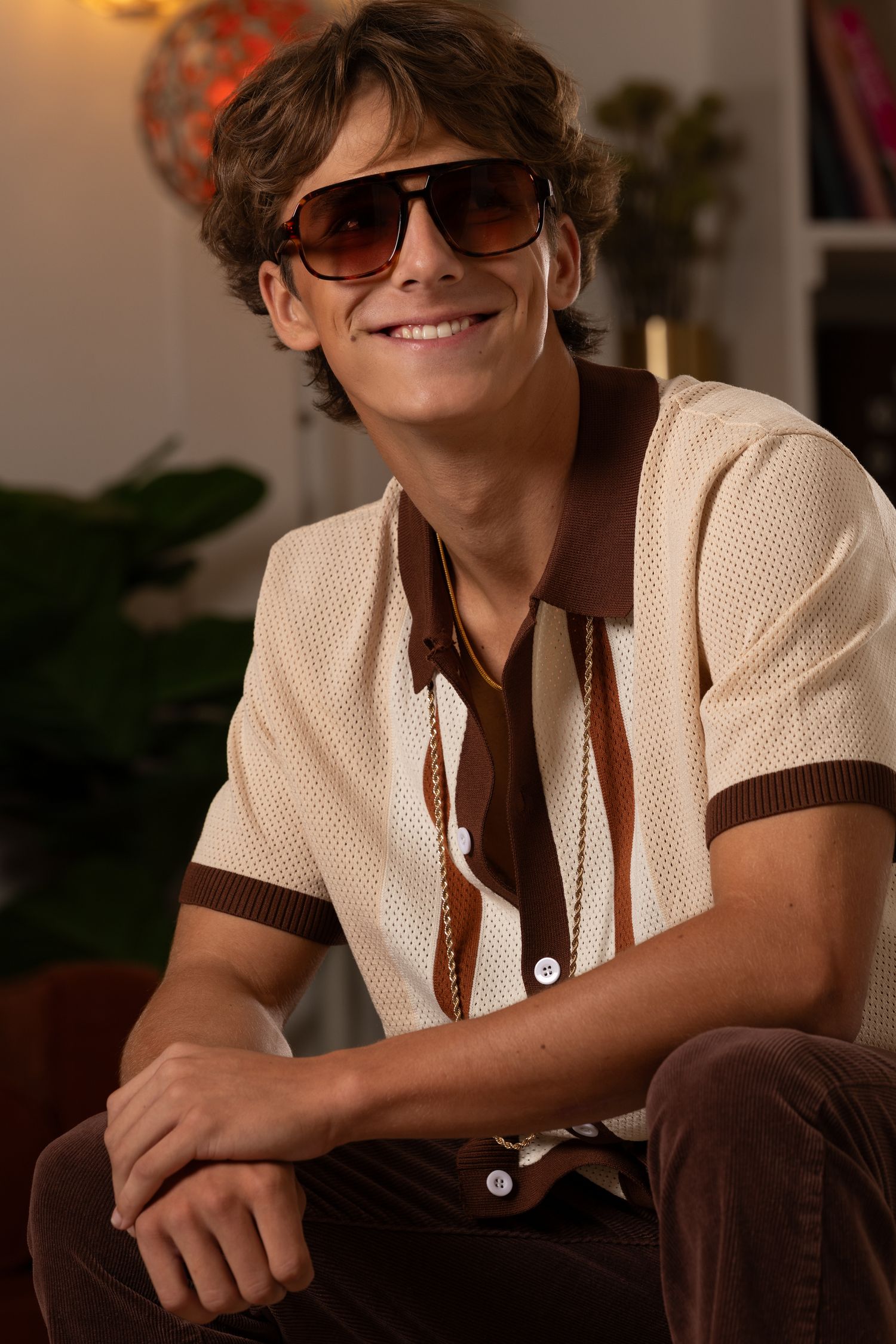Smiling young man wearing brown aviator sunglasses and a cream knit polo shirt with brown trim, seated indoors.