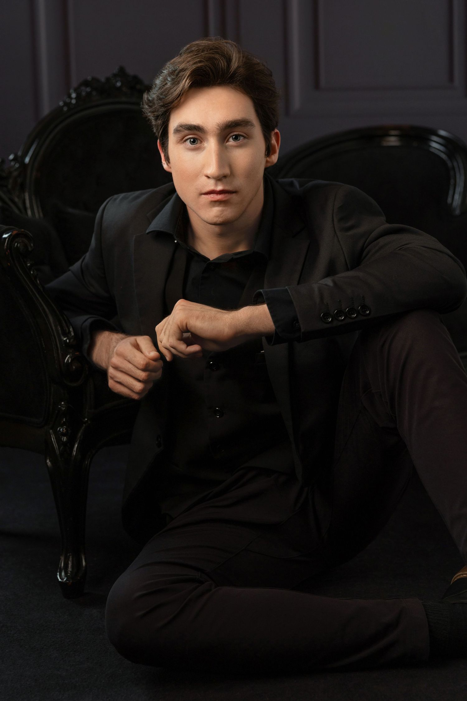 Young man in all-black suit sitting on floor leaning against ornate dark chair in dramatic studio portrait.