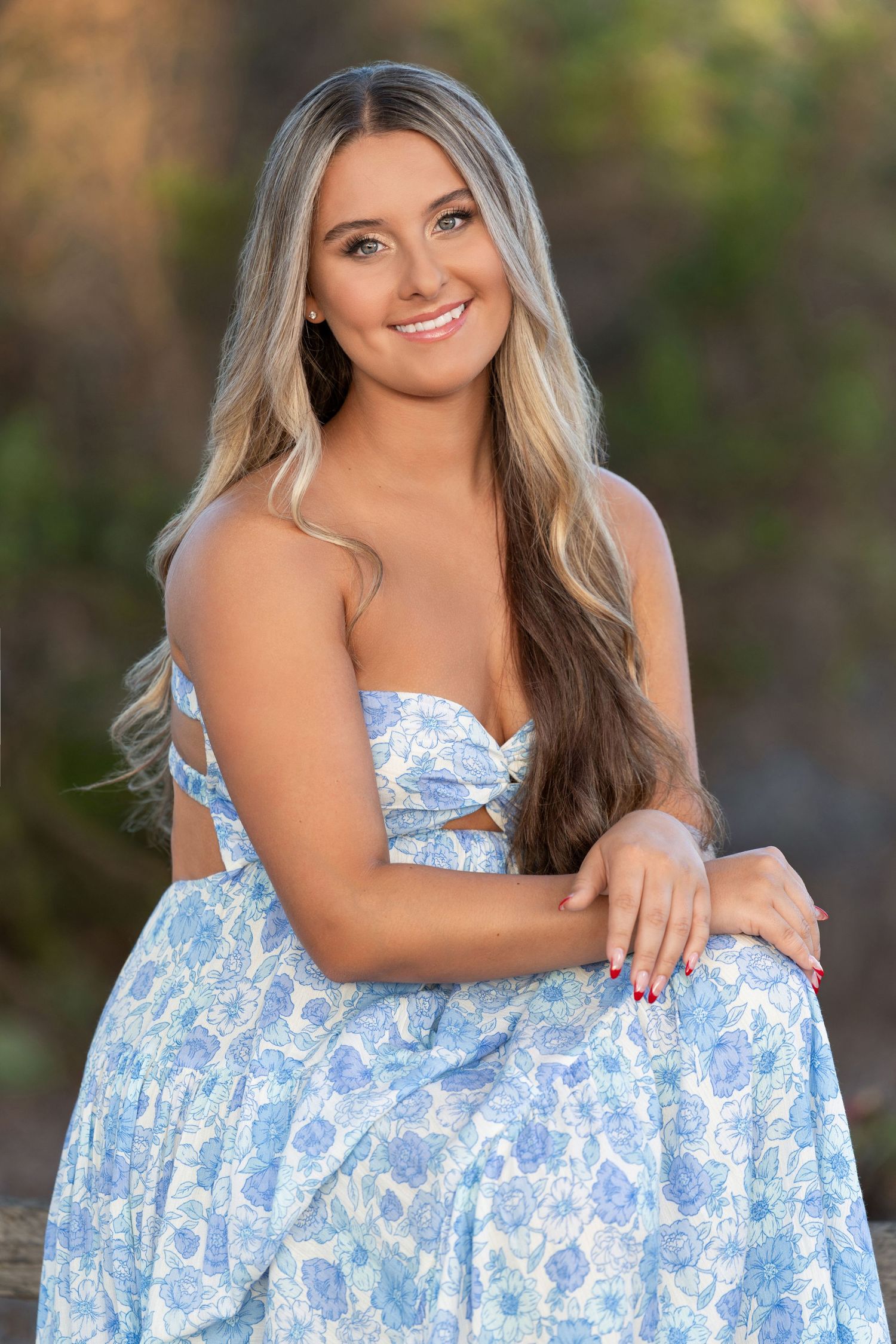 Young woman with long blonde hair smiling outdoors in a blue floral strapless dress.