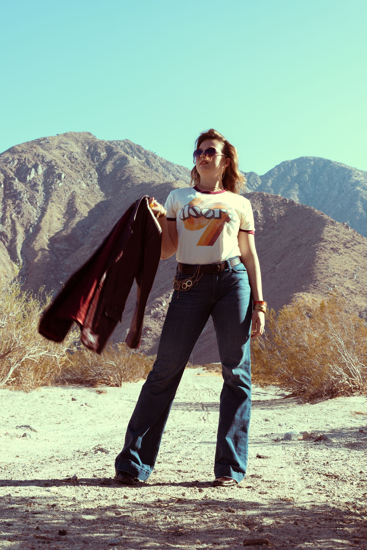 Woman in retro graphic tee, flare jeans, and sunglasses poses in desert landscape with mountains backdrop.
