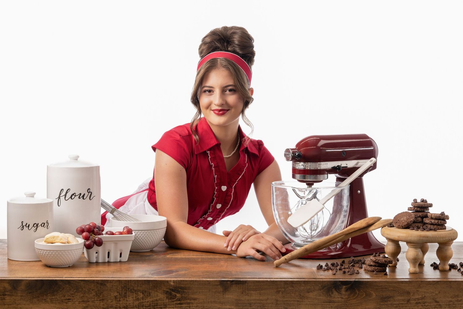 Smiling woman in red dress poses at wooden table with KitchenAid mixer, flour, sugar canisters, and baking ingredients.