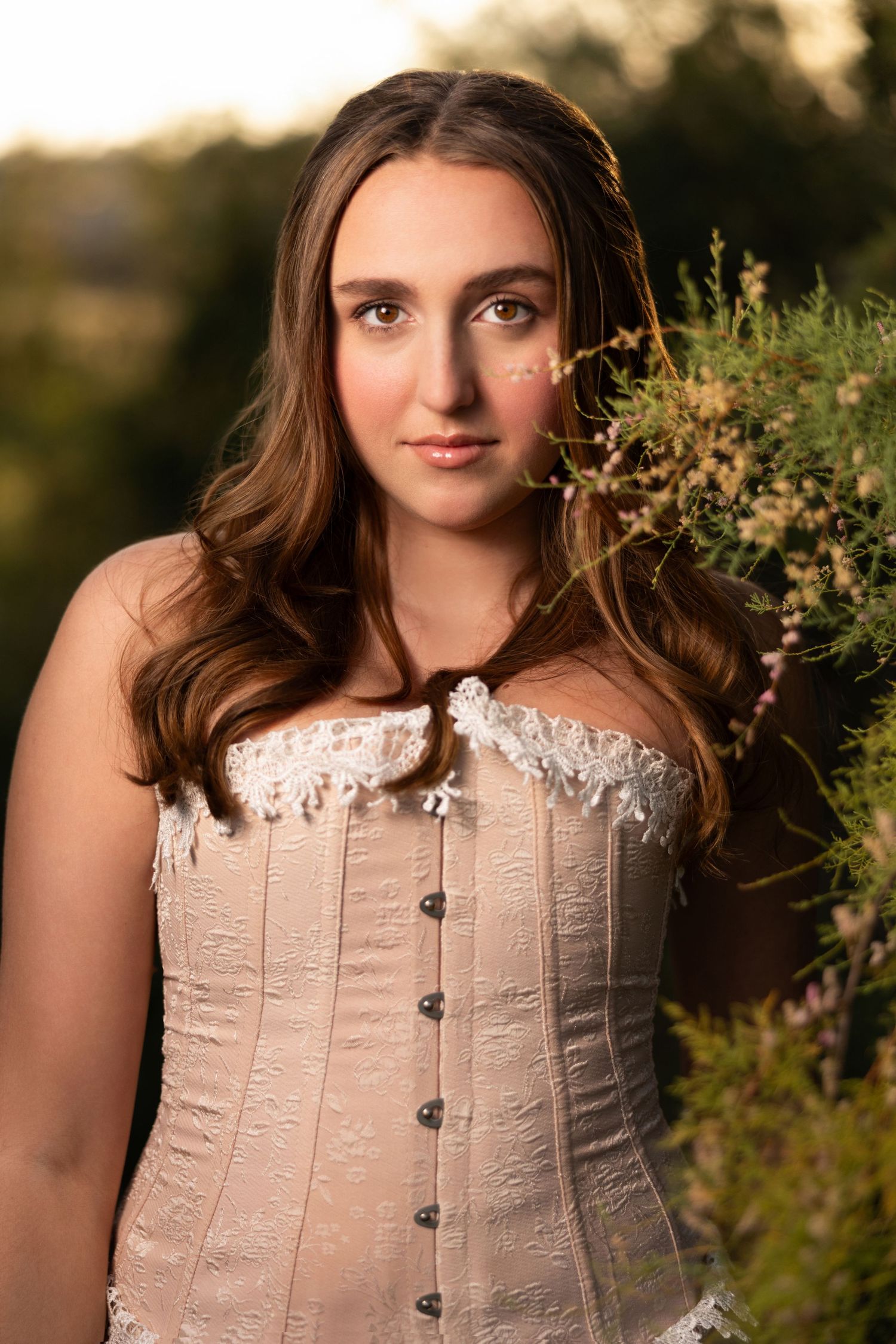 Young woman in a vintage cream corset poses outdoors at golden hour near wildflowers.