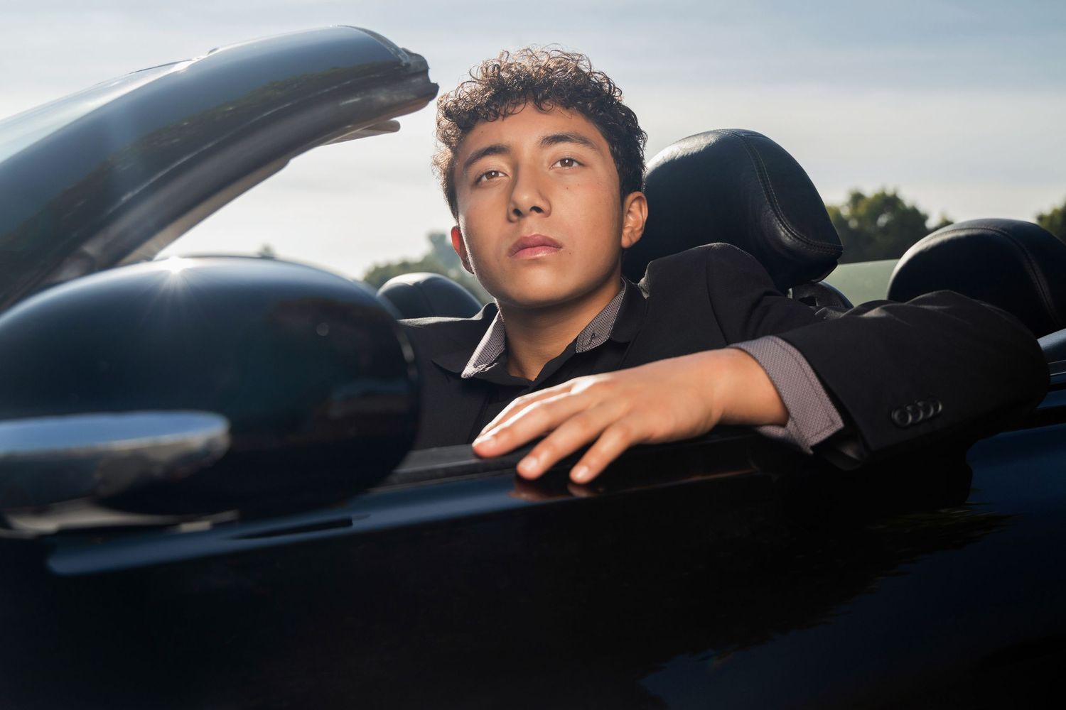 Young man in a suit sitting in a black convertible car, looking confidently at the camera.