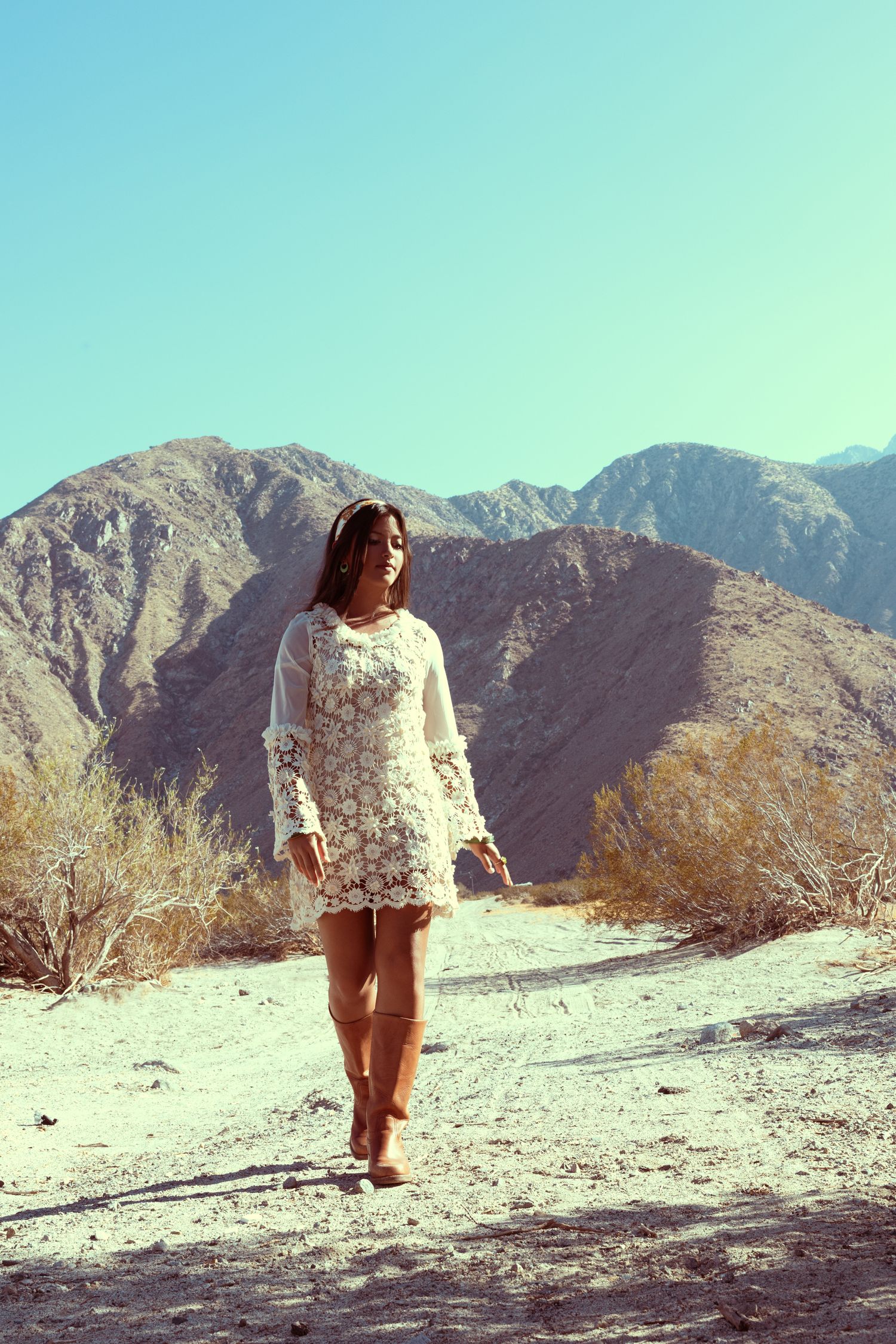 Woman in white lace dress and boots walks through desert landscape with mountains and clear blue sky.