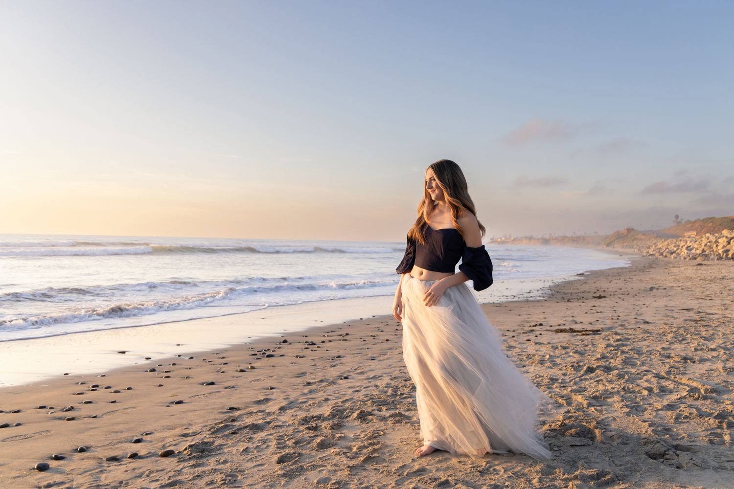 Woman in elegant white skirt and dark top poses on sandy beach at sunset with ocean waves in background.