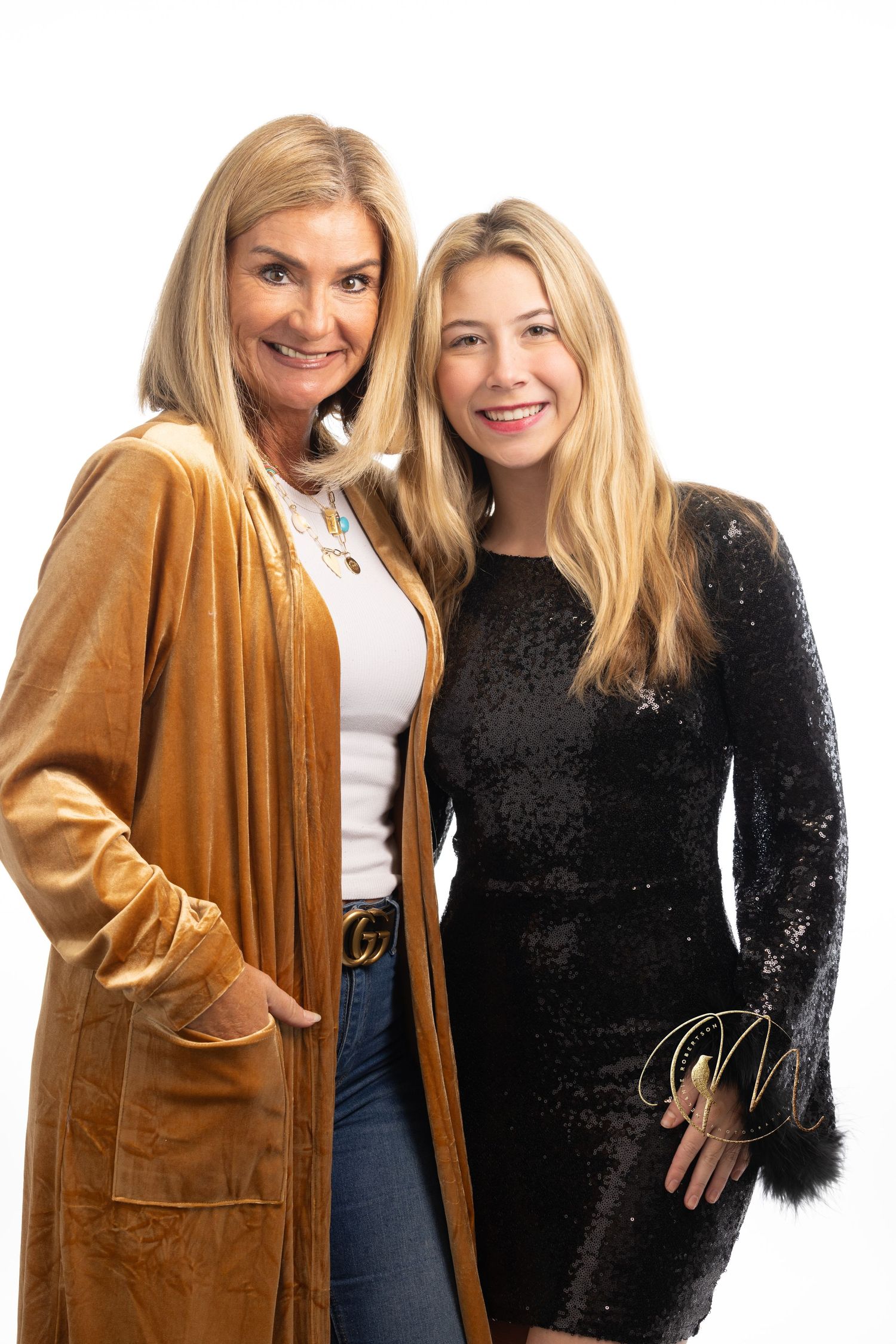 Two smiling blonde women pose together, one in a velvet caramel cardigan with jeans, the other in a black sequin mini dress.