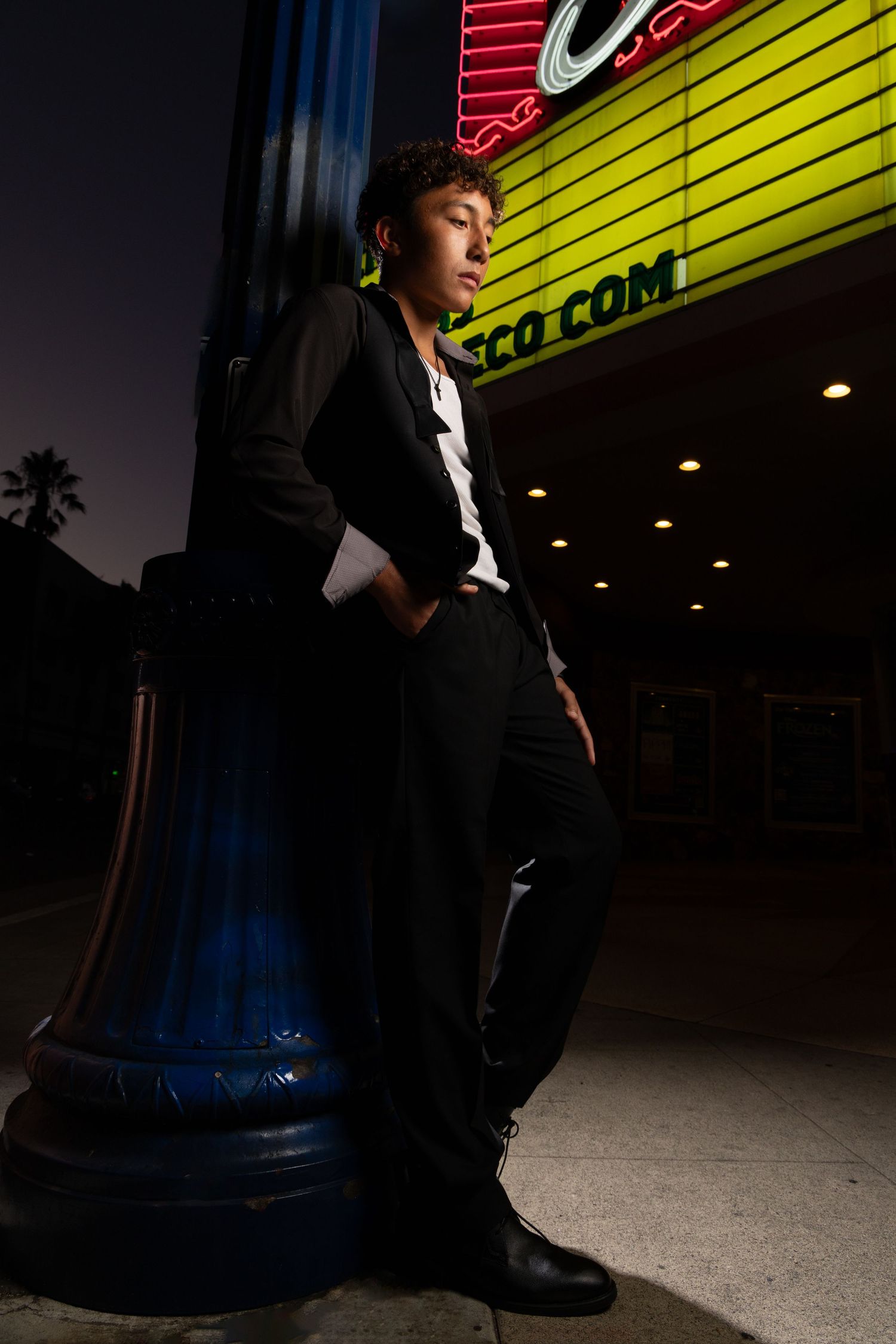 Young man in black suit leaning against a blue pillar outside a neon-lit theater at night.