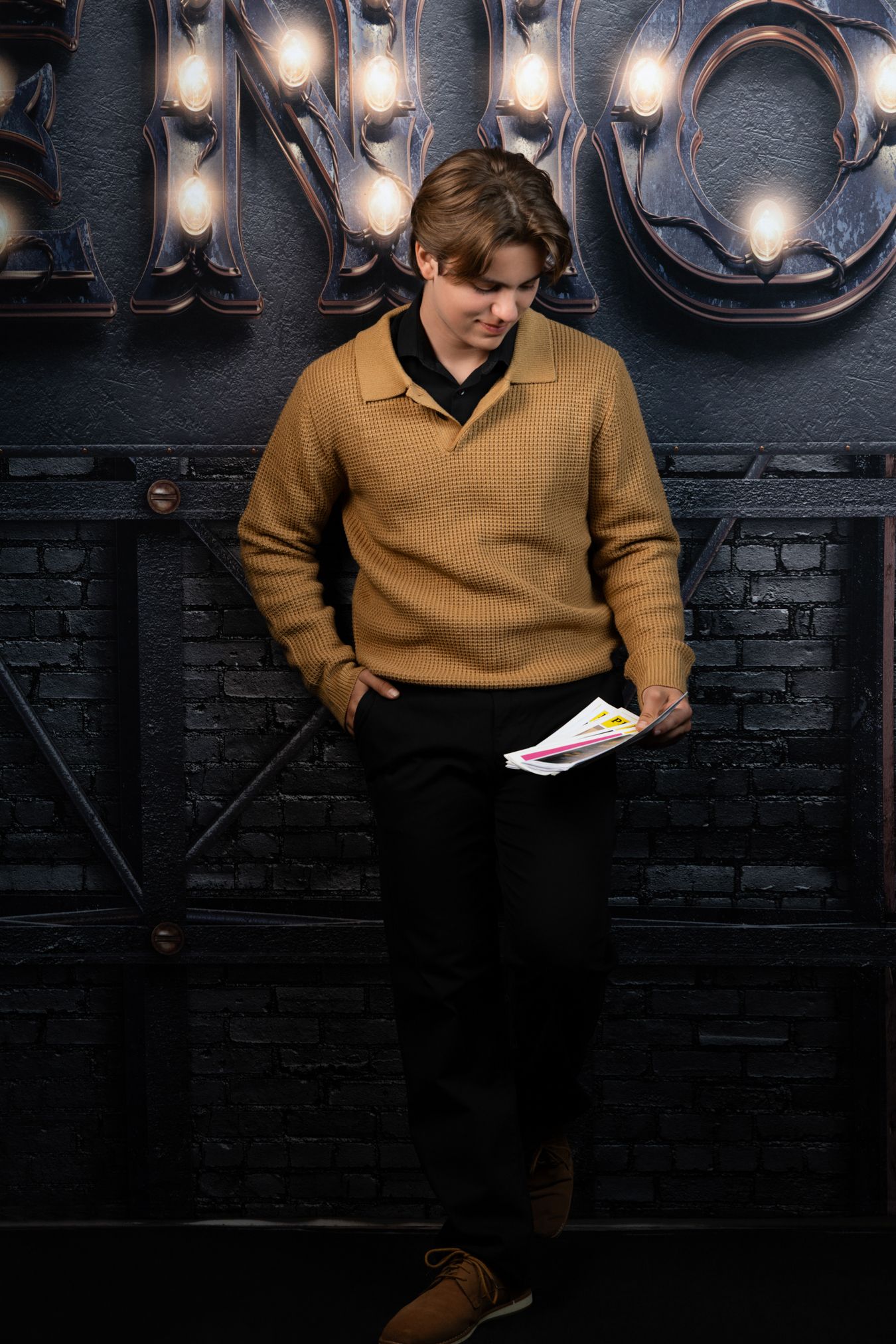 Young man in tan sweater holding papers, leaning against dark brick wall with marquee lights.
