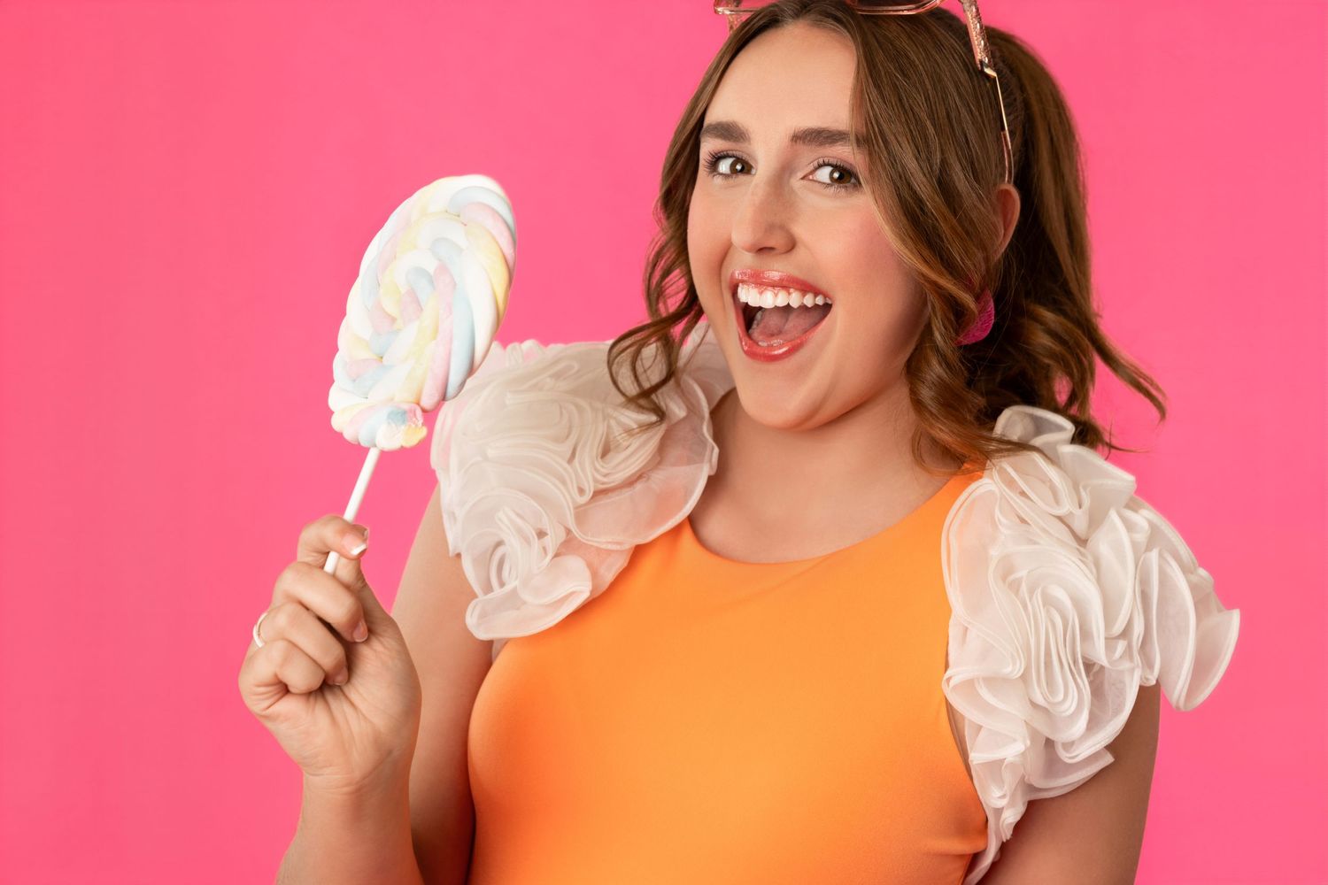 Woman in orange top holding large lollipop, smiling against pink background.