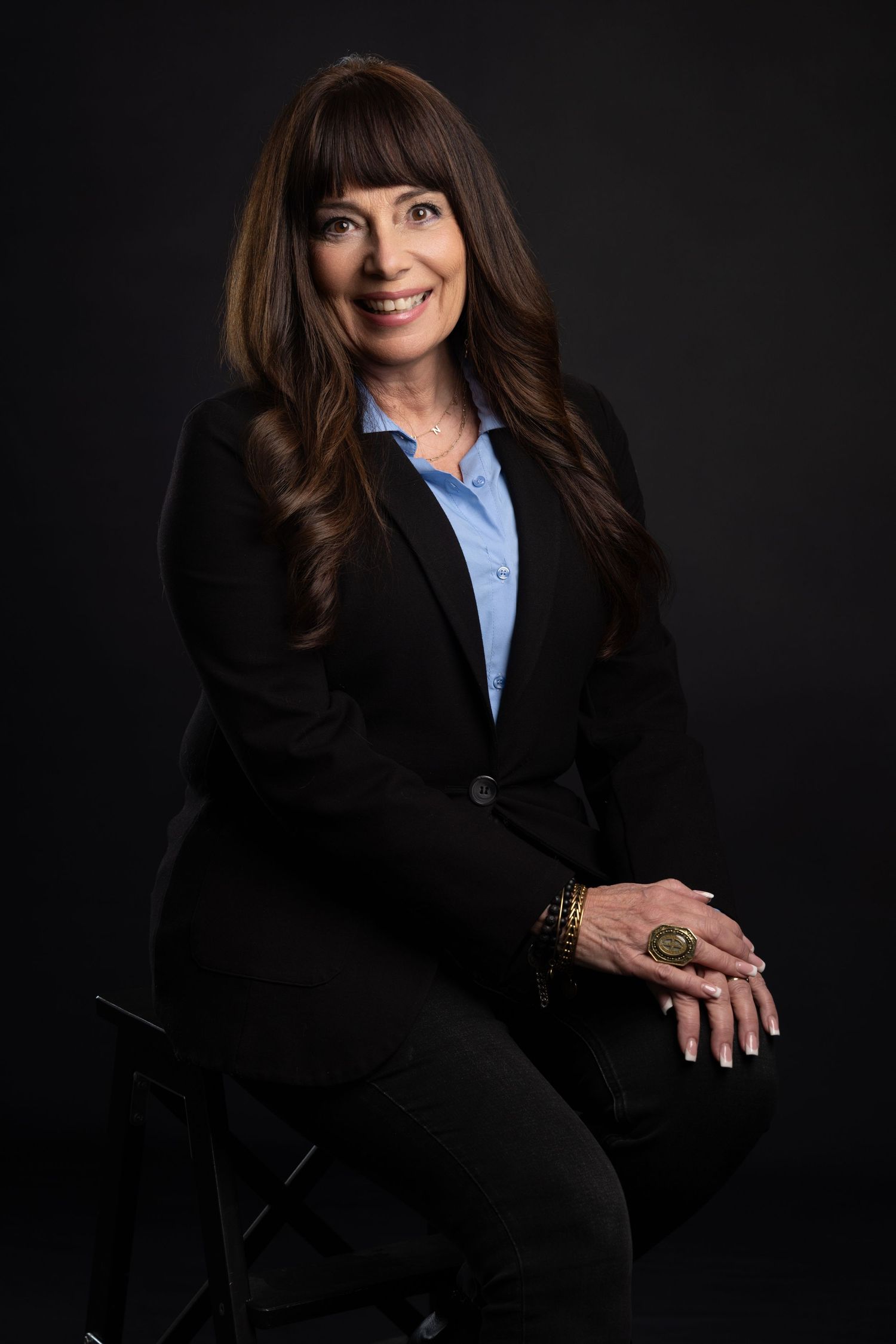 Professional woman in black blazer and blue shirt seated confidently against dark background, smiling warmly.